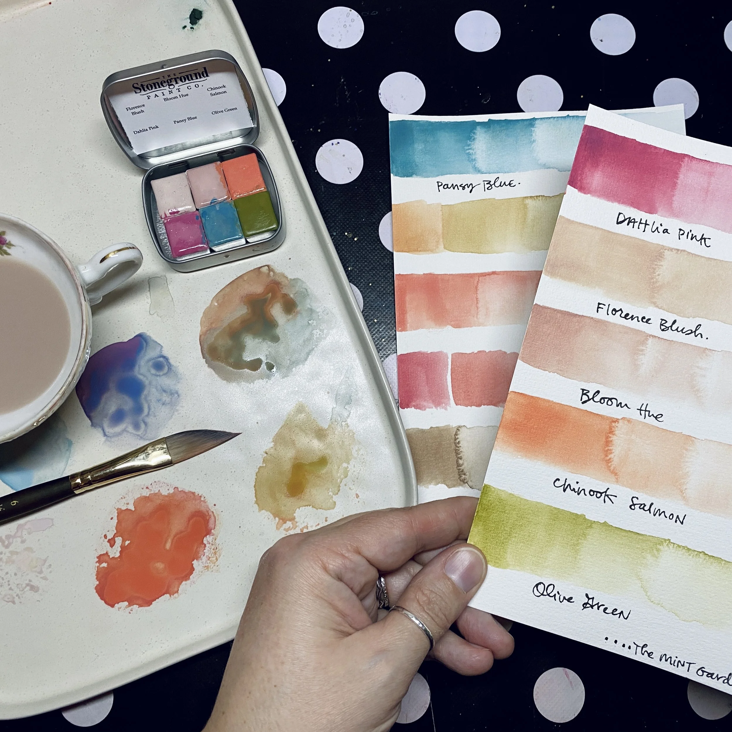 A hand holding a color palette with watercolor paint swatches and handwritten labels, surrounded by watercolor swatches and a cup of tea or coffee on a polka dot table.