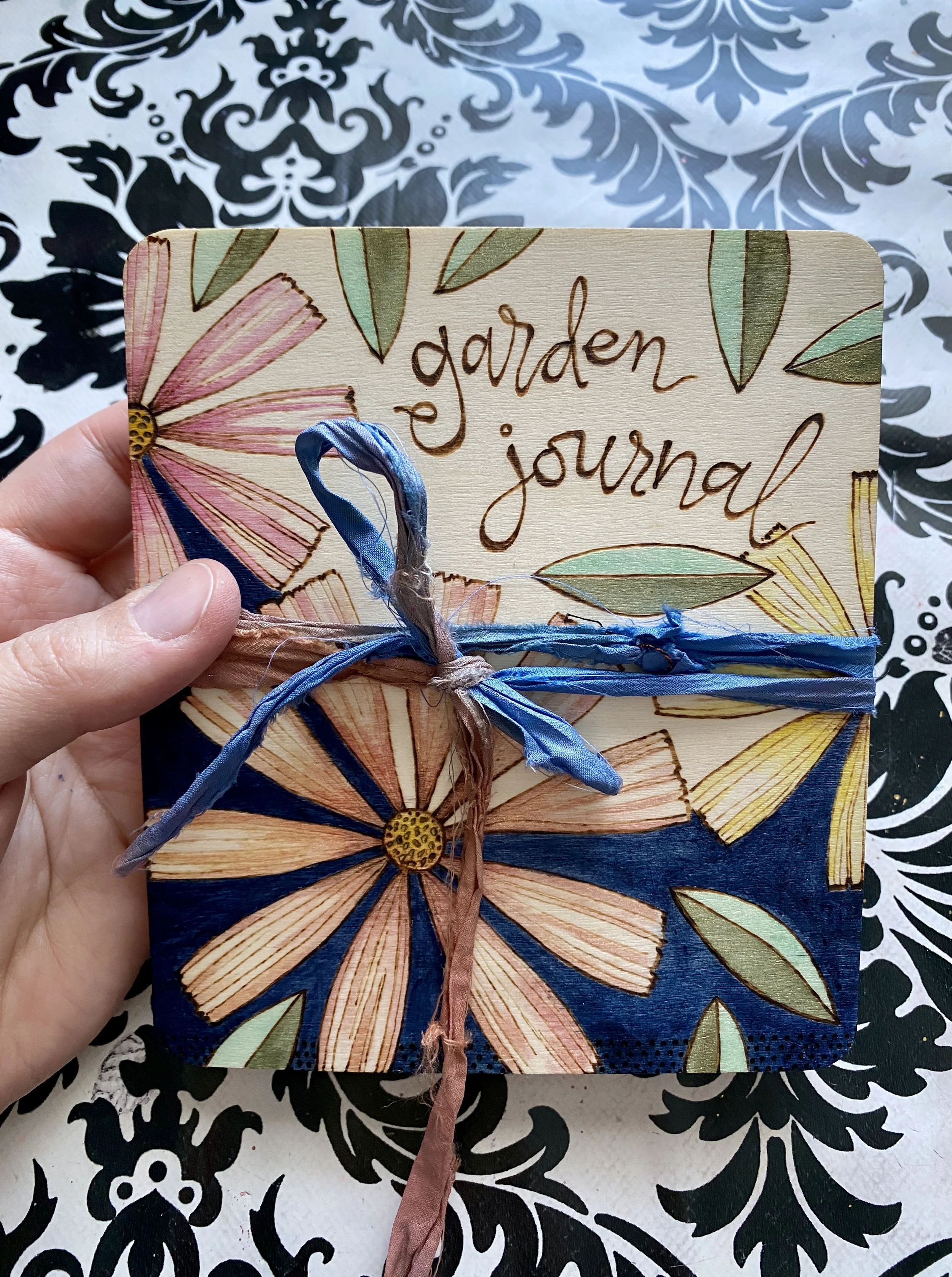 A hand holding a small decorative journal with a floral cover design labeled "garden journal" tied with pink and blue ribbons, against a black and white floral patterned background.