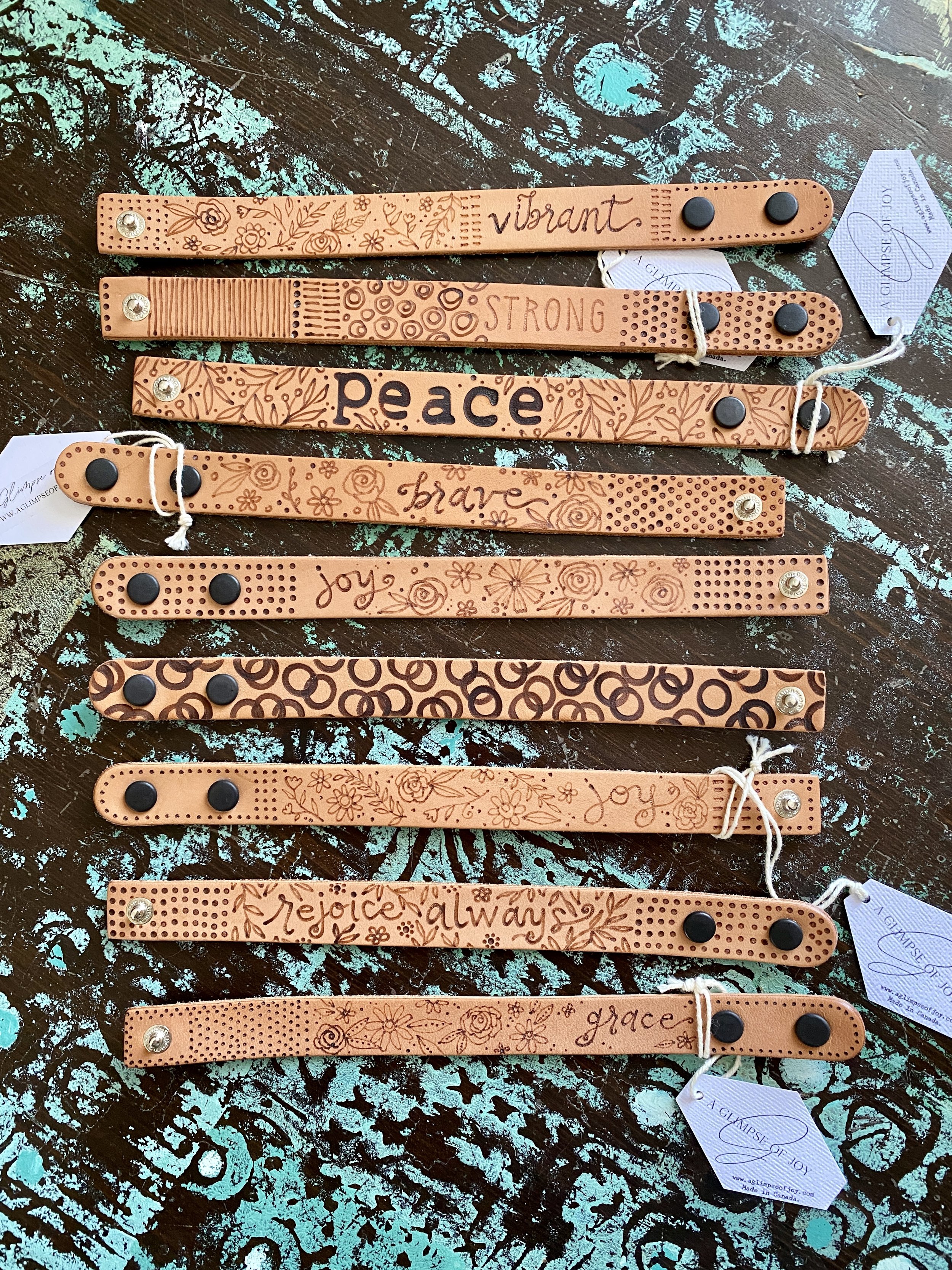 Decorative leather wristbands with inspirational words and floral patterns, arranged on a rustic wooden surface.