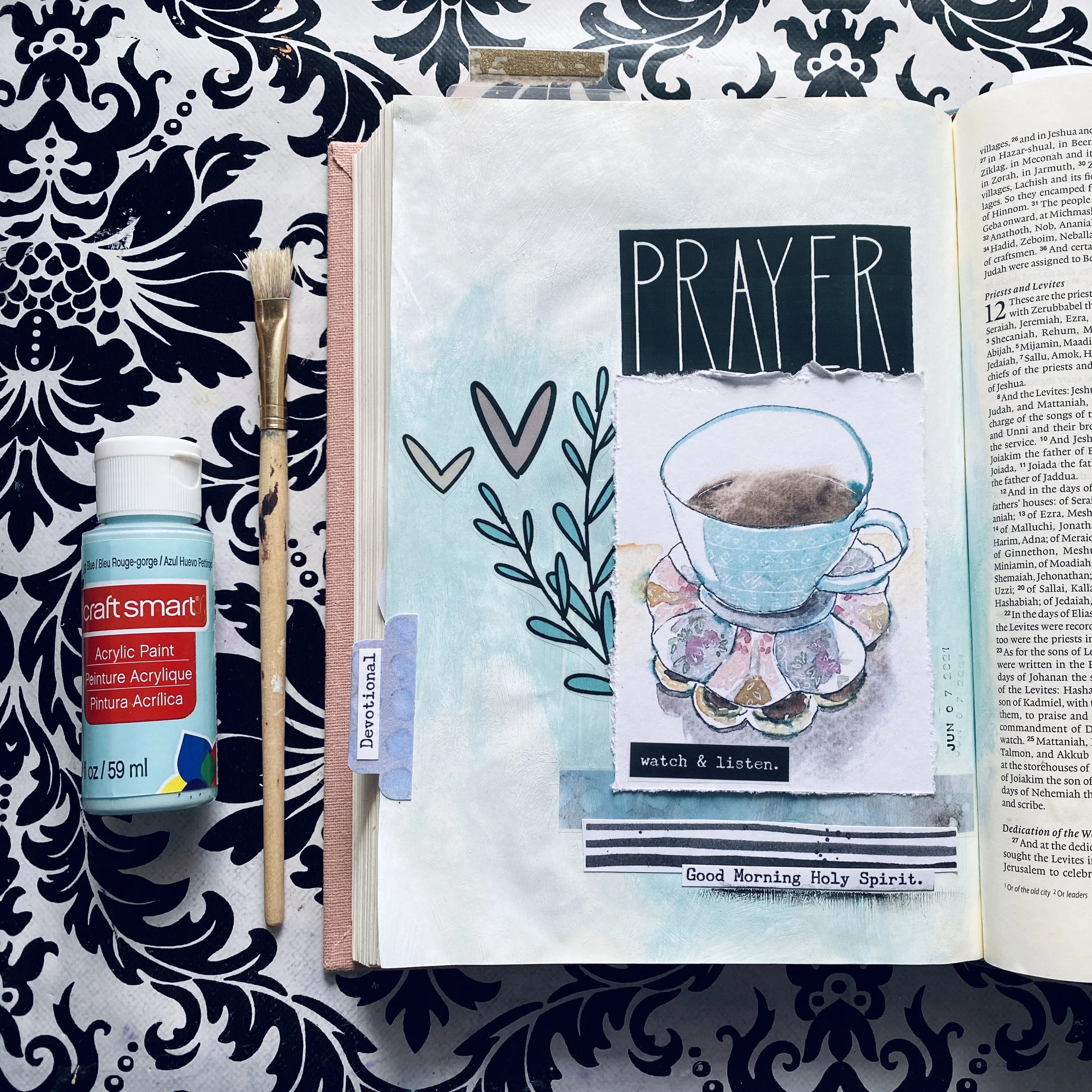 An open journal with a mixed media art piece on the right page, featuring a watercolor cup of coffee, surrounded by words 'watch & listen.' and 'Good Morning Holy Spirit.' On the left, there is a bottle of acrylic paint, a small paintbrush, and a sheet of decorative paper underneath. The journal is placed on a black and white floral patterned fabric.