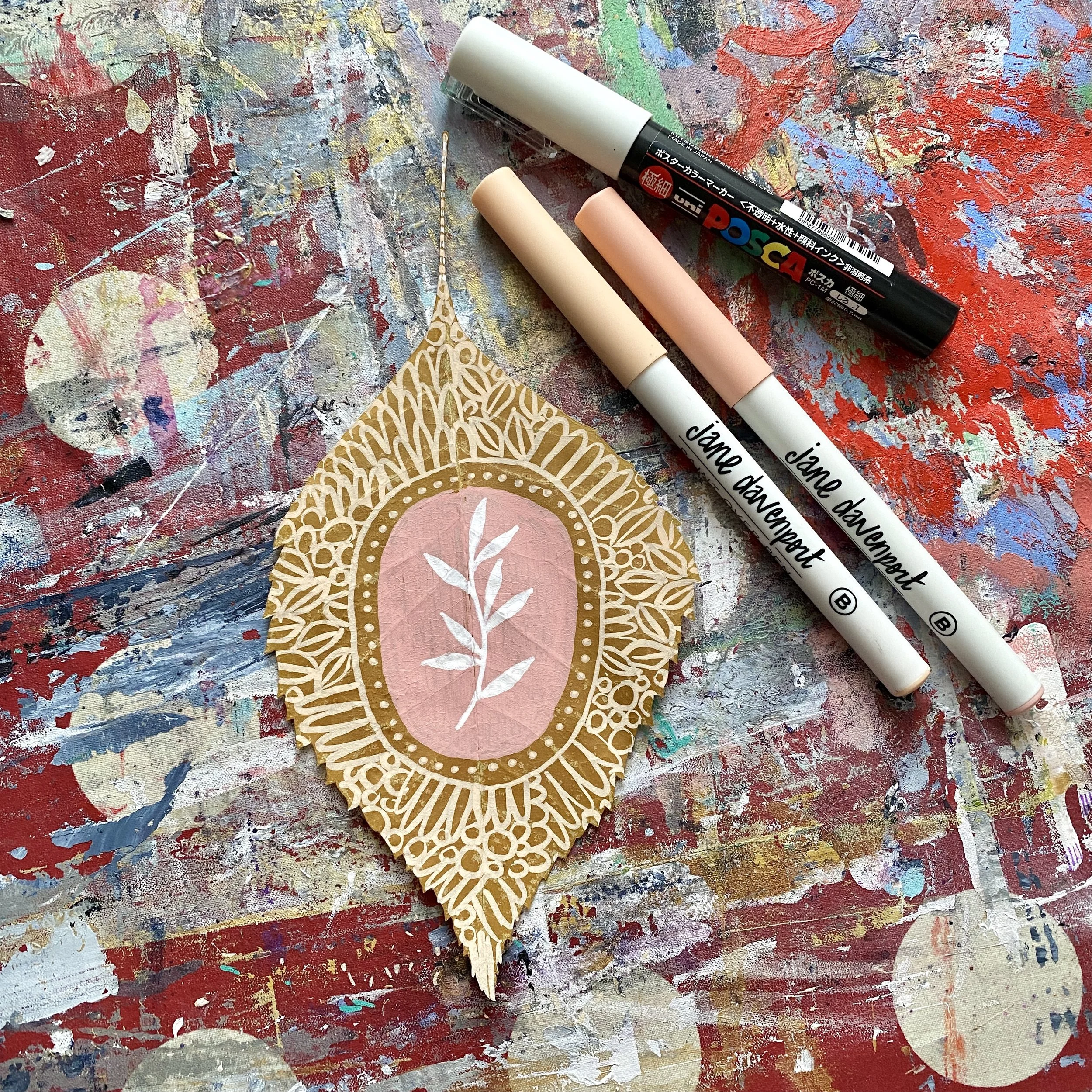 Decorative paper cutout with intricate gold patterns and a pink center featuring white leaf illustrations, surrounded by artist markers on a colorful, paint-splattered surface.