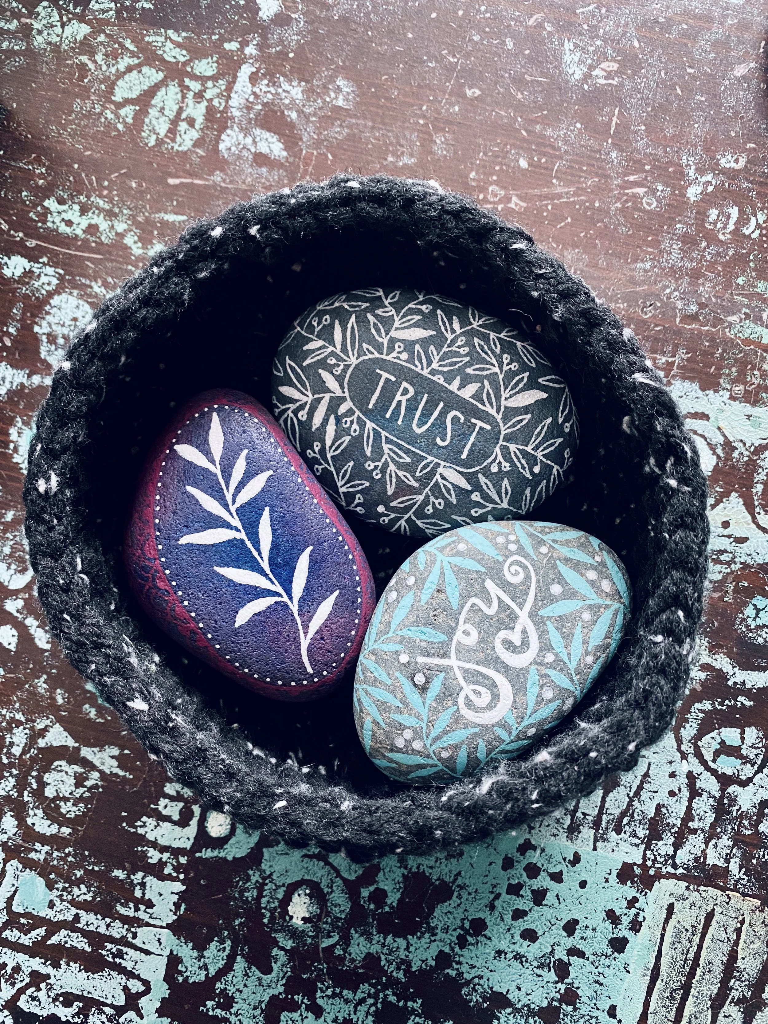 Decorated rocks with painted designs and words inside a black basket, placed on a distressed wooden surface.