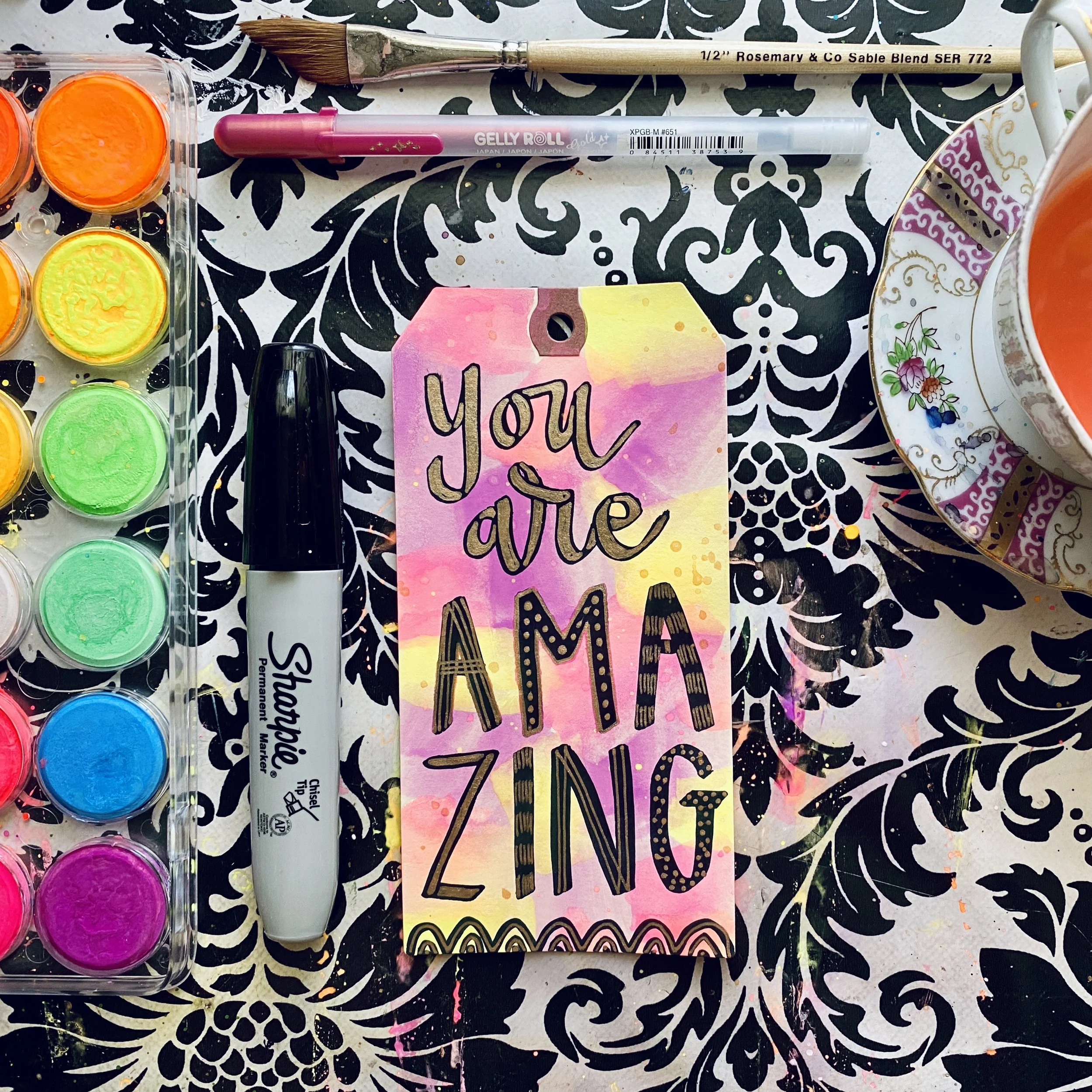 Colorful painted tag with the message 'You are amazing', surrounded by paint set, marker, and tea cup on a patterned tablecloth.
