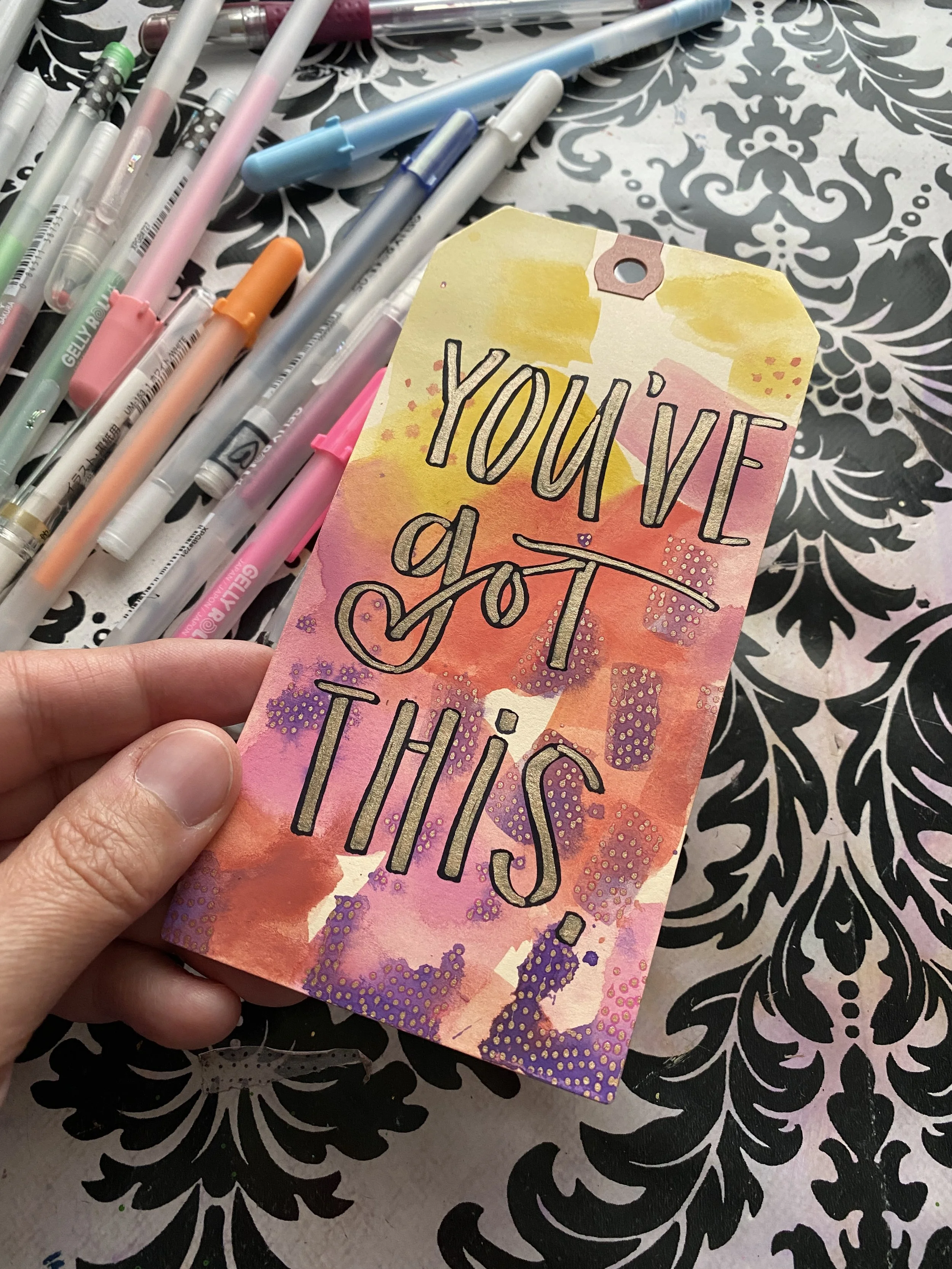 A colorful hand-painted tag with the motivational quote 'You’ve got this' in black outlined letters, held over a black and white decorative patterned background, with a table of assorted pastel-colored pens nearby.