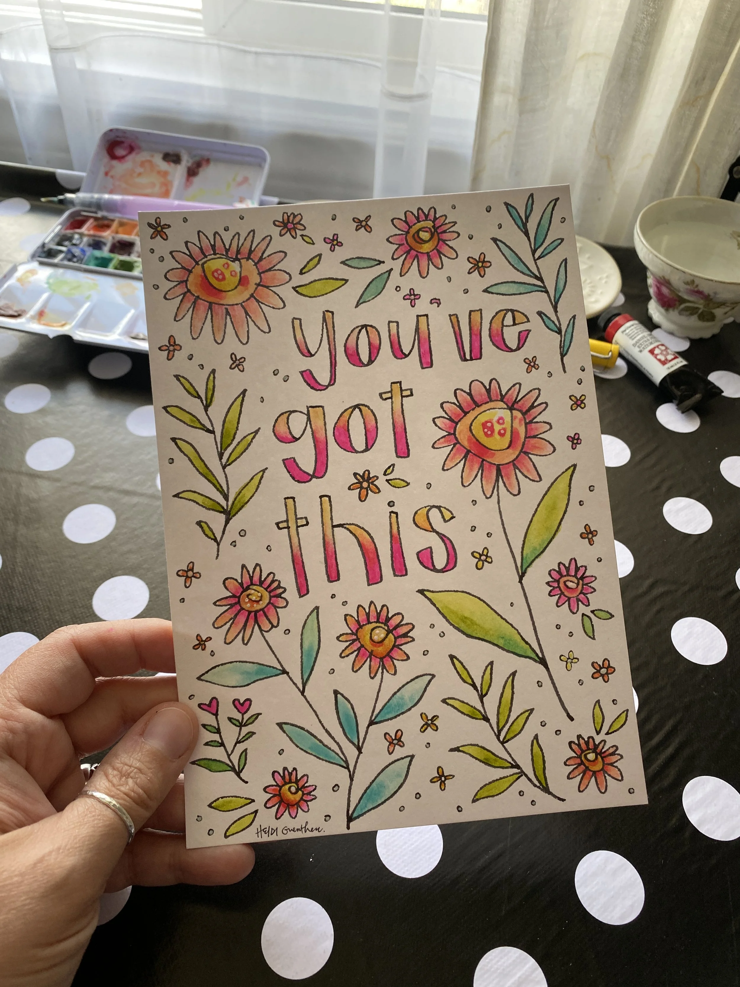 Hand holding a colorful card with the phrase 'you've got this' surrounded by pink, orange, and yellow flowers and green leaves, on a black table with white polka dots, art supplies, and a bowl in the background.