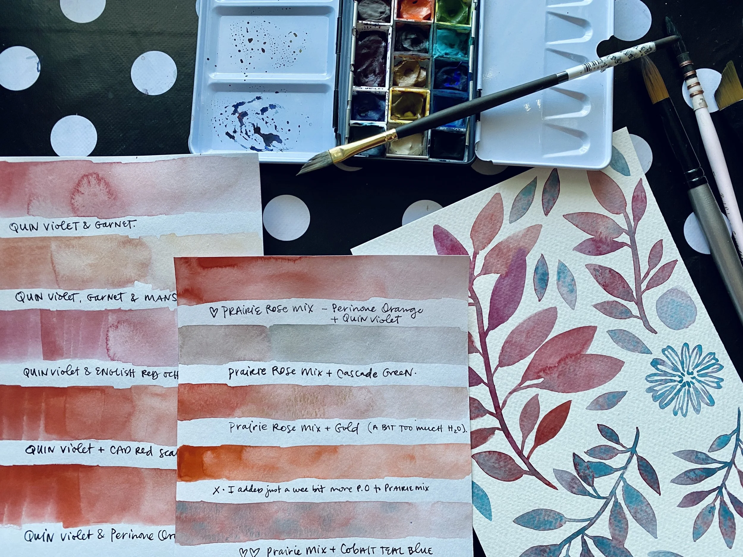 A watercolor painting setup featuring a palette of watercolors, paintbrushes, and two sheets of paper with watercolor swatches and notes. One paper includes color swatches labeled with shades of quinn violet, garnet, and other colors, while the other