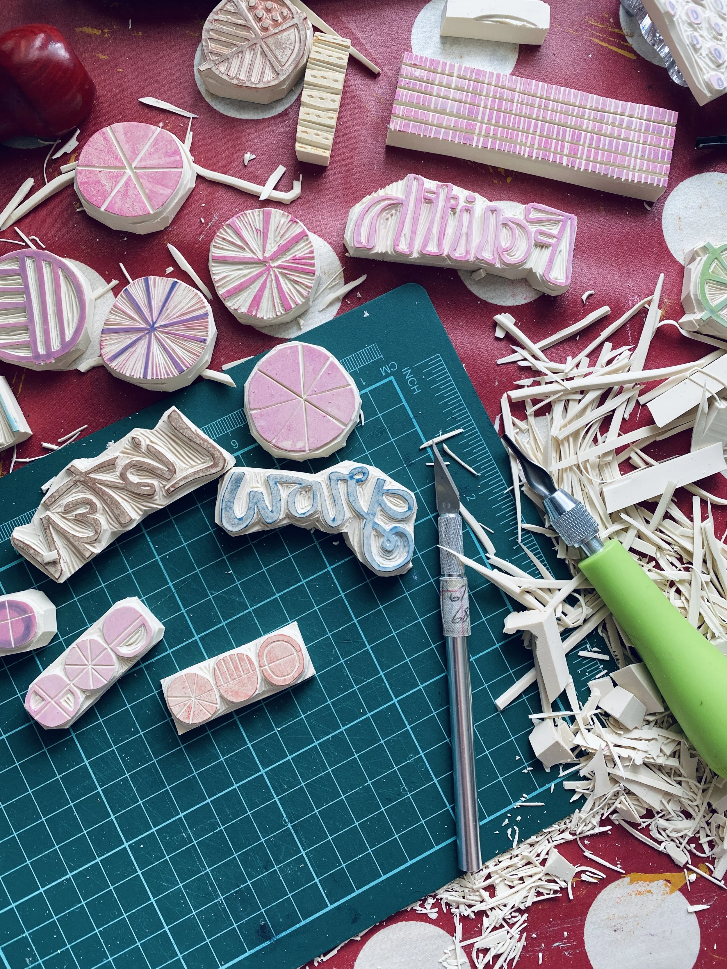 Colorful rubber stamps, carving tools, and paper shavings on a cutting mat, used for making custom rubber stamps.