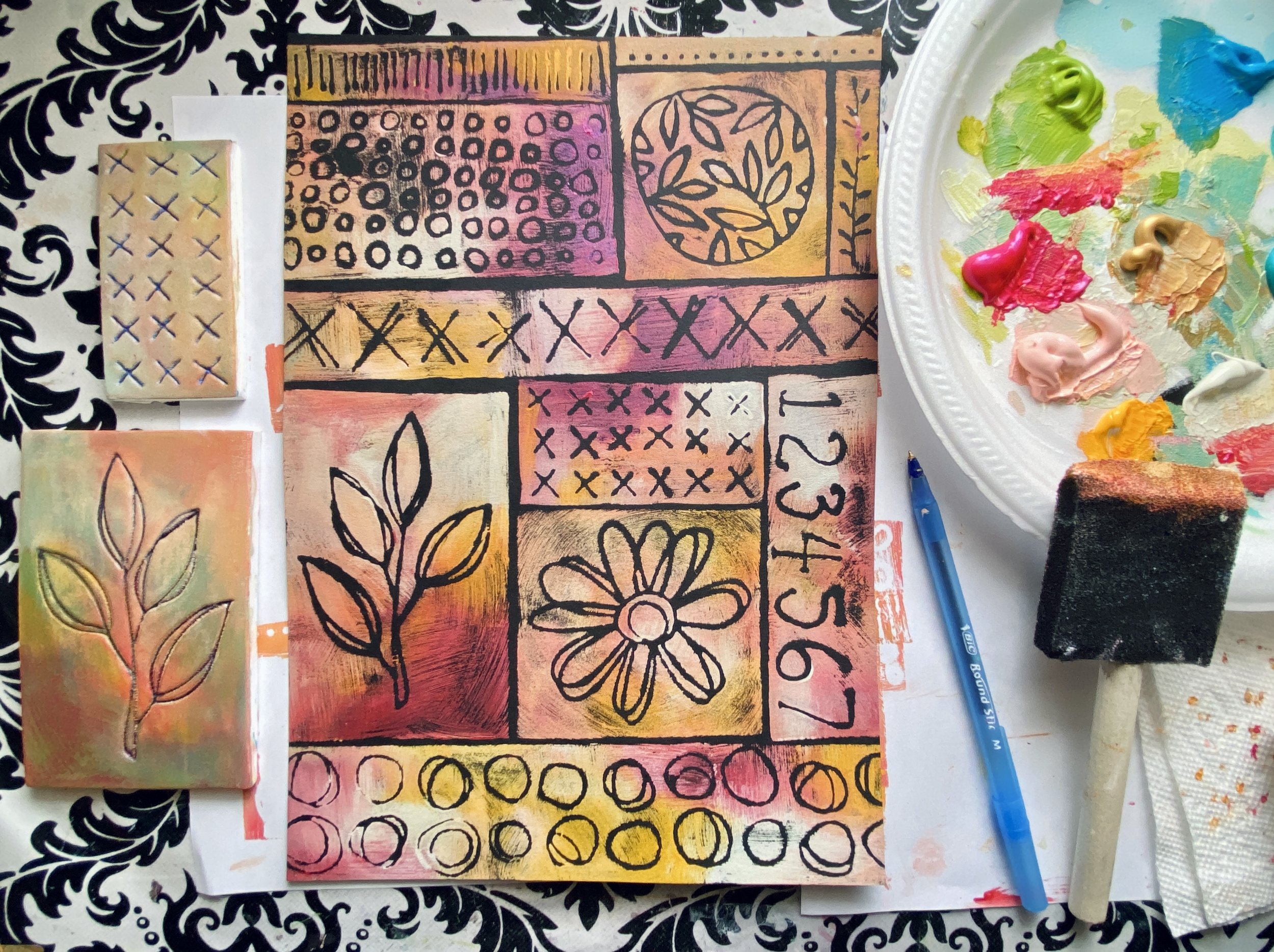 A mixed media art project on paper with floral and geometric patterns, painted in pink, yellow, orange, purple, and black. Art supplies include paint on a plate, a paint roller, and a blue pen, all on a black and white patterned surface.
