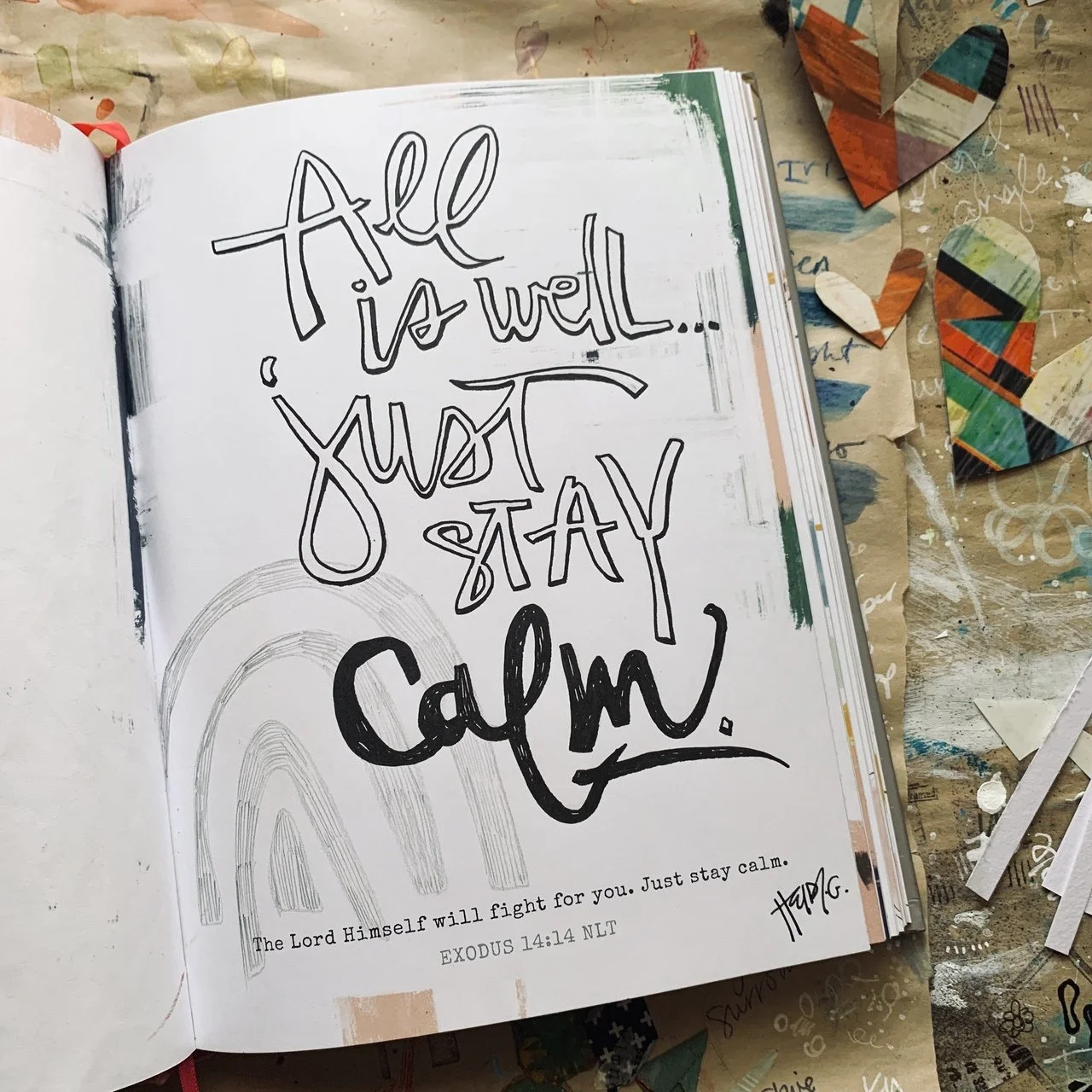 An open Bible with handwritten text saying "All is well... just stay calm" and a Bible verse from Exodus 14:14. The background has scattered paper hearts and arts and crafts supplies.