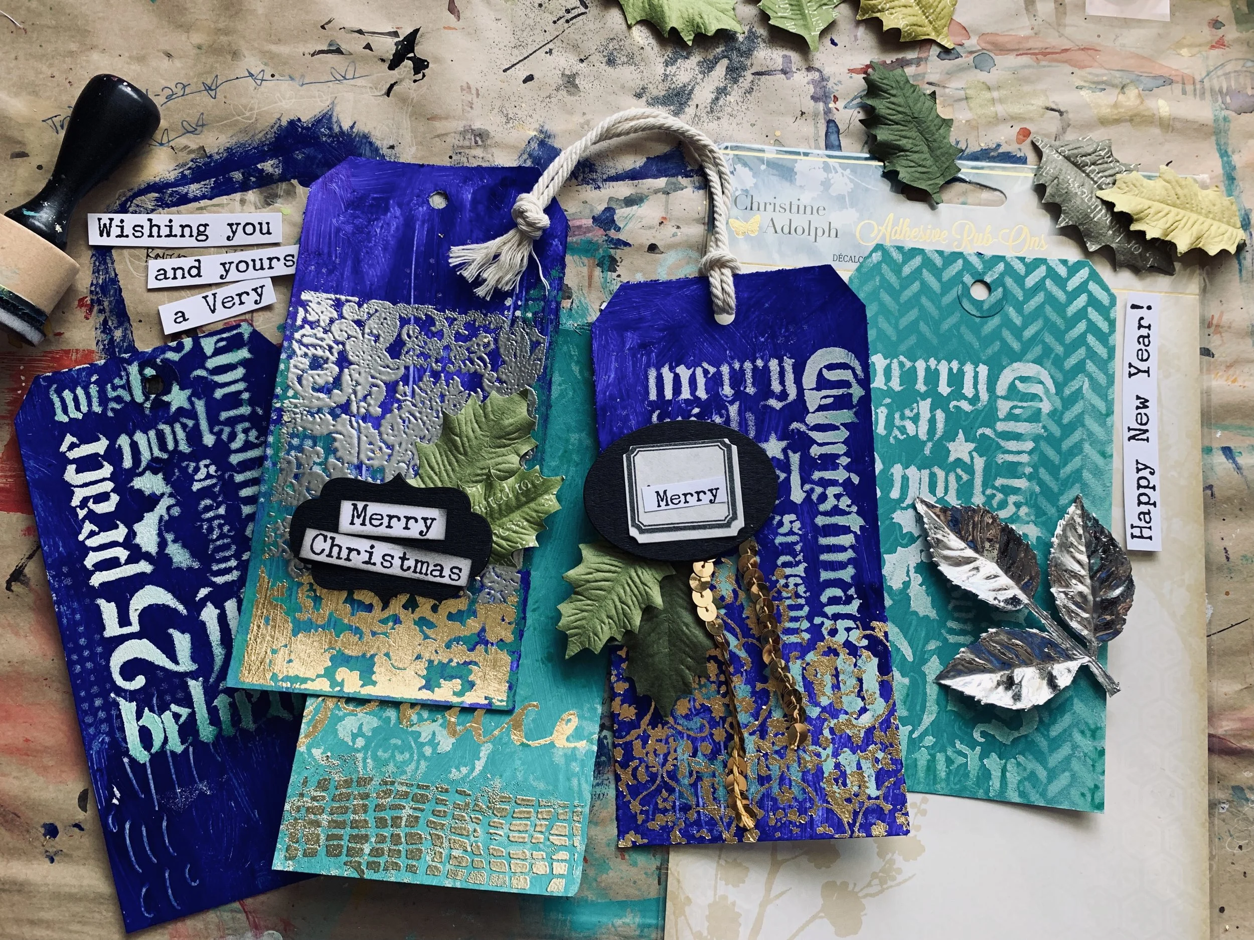 An assortment of decorated Christmas tags with varying shades of blue, gold, and silver, featuring Christmas-themed stamps and embellishments. Some tags have Christmas messages like "Merry Christmas" and "Happy New Year!" with tags, leaves, and sequins scattered on a paint-splattered surface.
