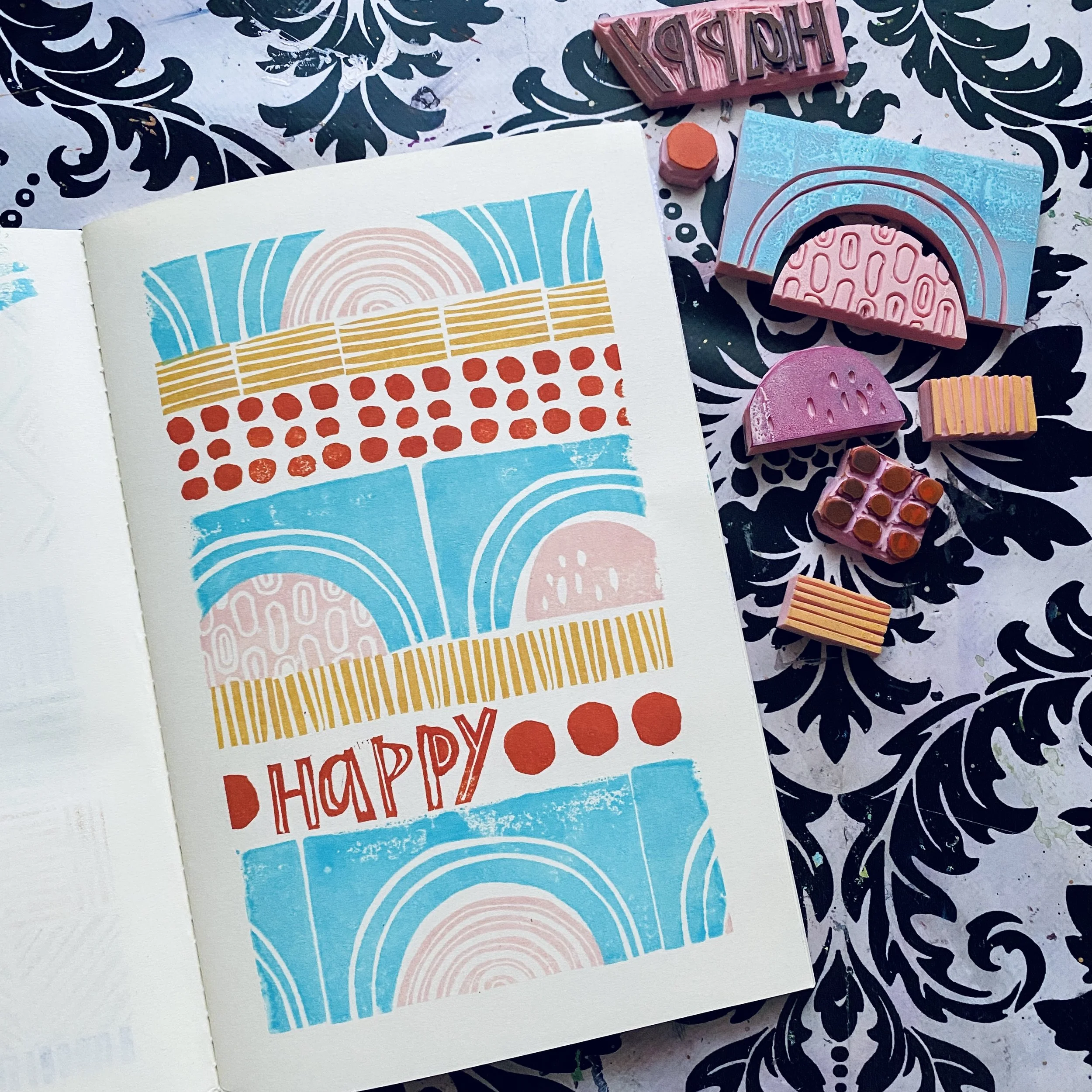A colorful birthday card with abstract geometric patterns and the word "HAPPY" written on it. Surrounding the card are rubber stamps and ink pads in various colors, placed on a black and white floral patterned surface.