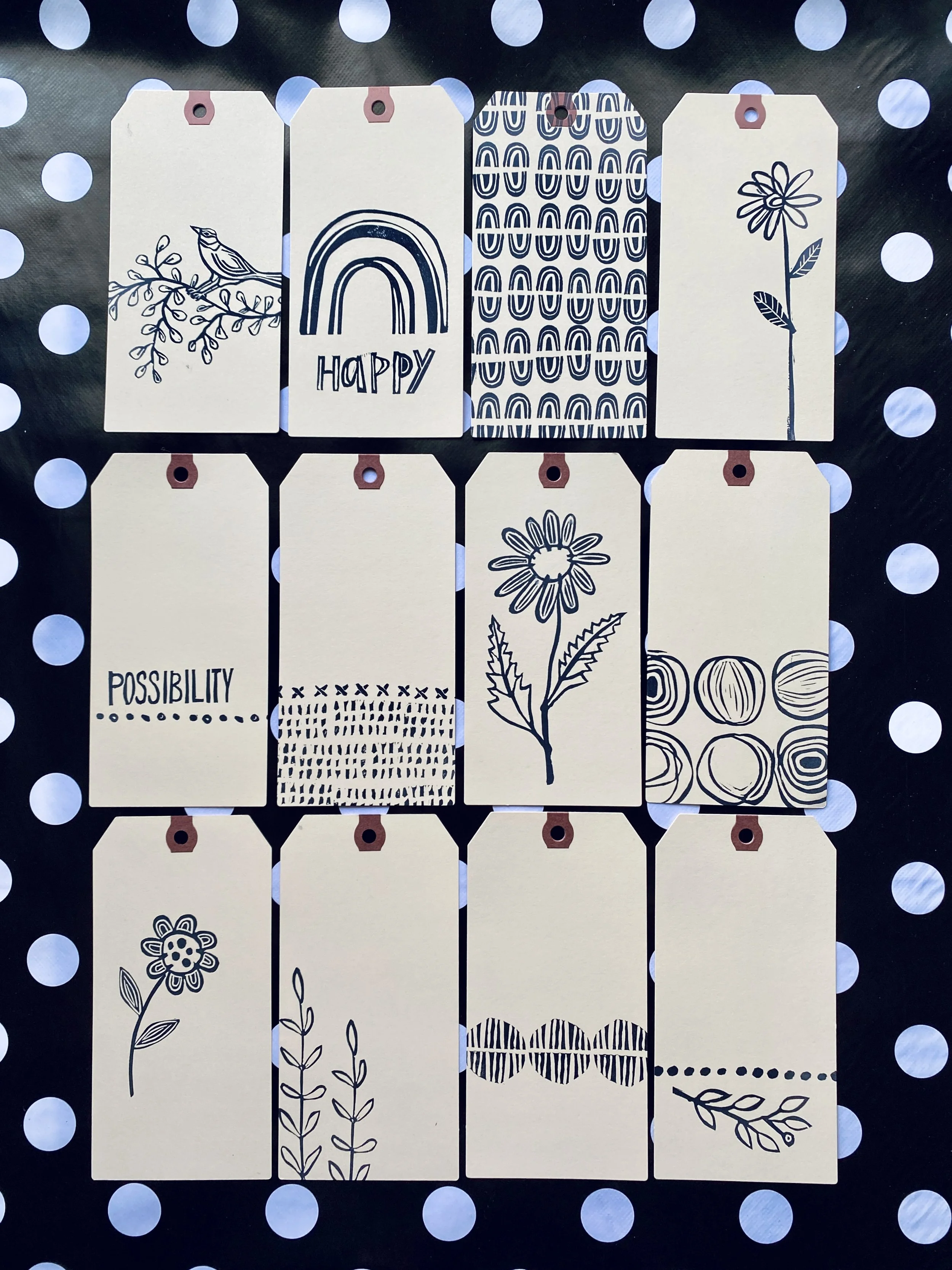 Set of 12 decorative kraft paper tags with black line drawings and text, arranged on black paper with white polka dots. Designs include a bird on a branch, a rainbow with the word 'HAPPY,' abstract patterns, a flower, and words like 'POSSIBILITY' wit