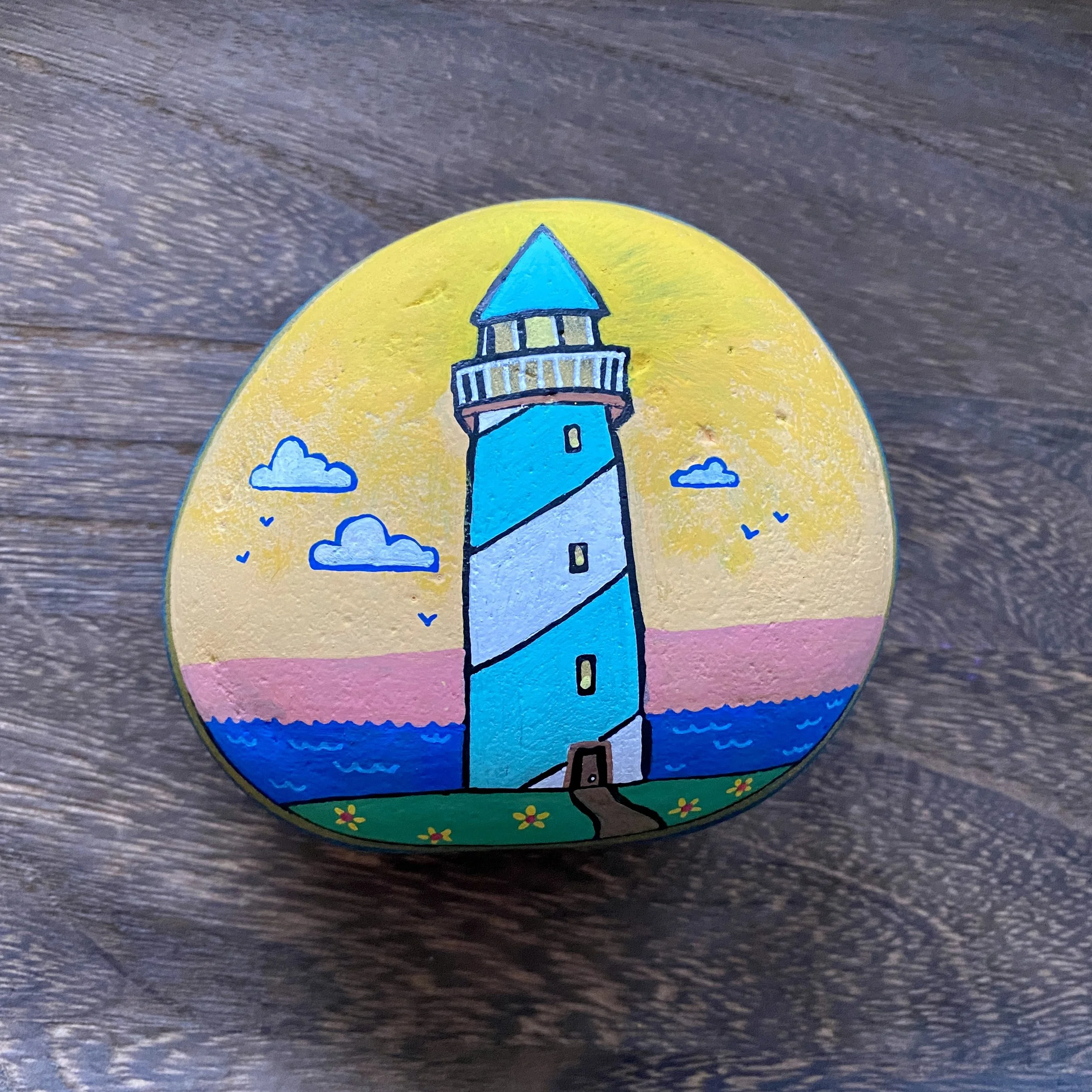 Hand-painted rock featuring a lighthouse by the water with a sunset sky, clouds, and flowers in the foreground.
