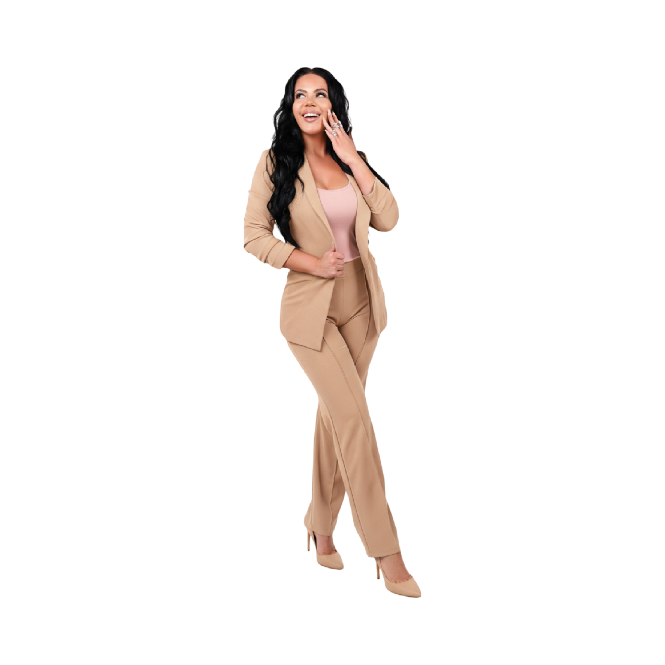 Woman with long dark hair wearing a beige pantsuit, light pink top, and beige high heels, smiling with one hand touching her face.