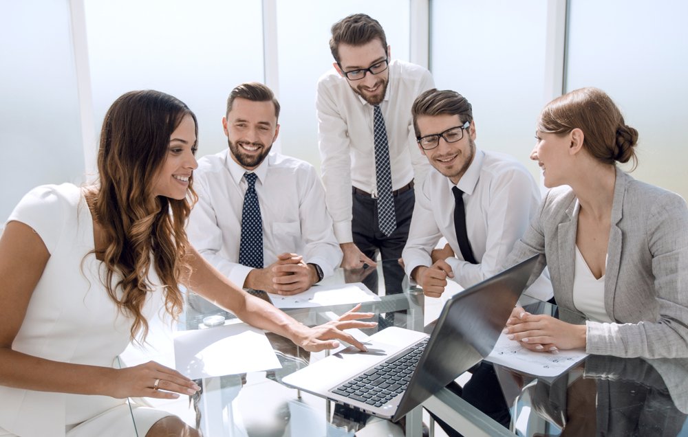 Grupo de personas en una reunión de negocios, todos sonrientes, alrededor de una mesa con una computadora portátil.