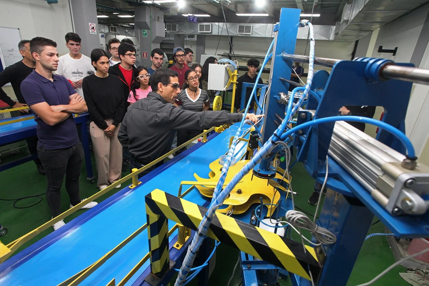 Grupo de personas observando una demostración en un laboratorio industrial con maquinaria azul y amarilla.