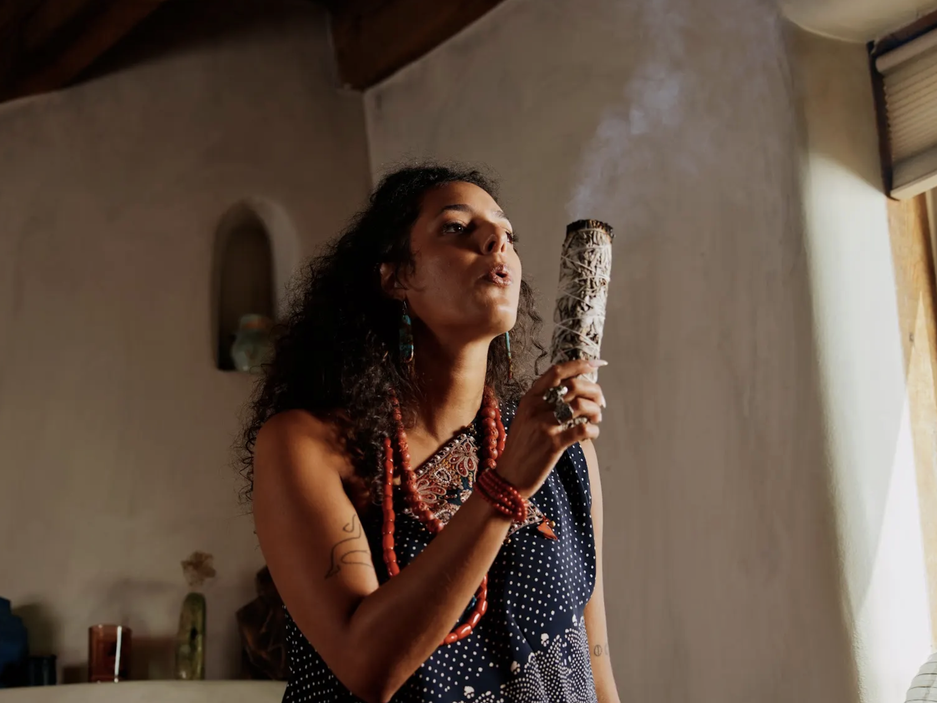 A woman with curly dark hair, wearing colorful jewelry and a polka-dot dress, holding a carved wooden object with smoke rising from it inside a room with a wall and window.