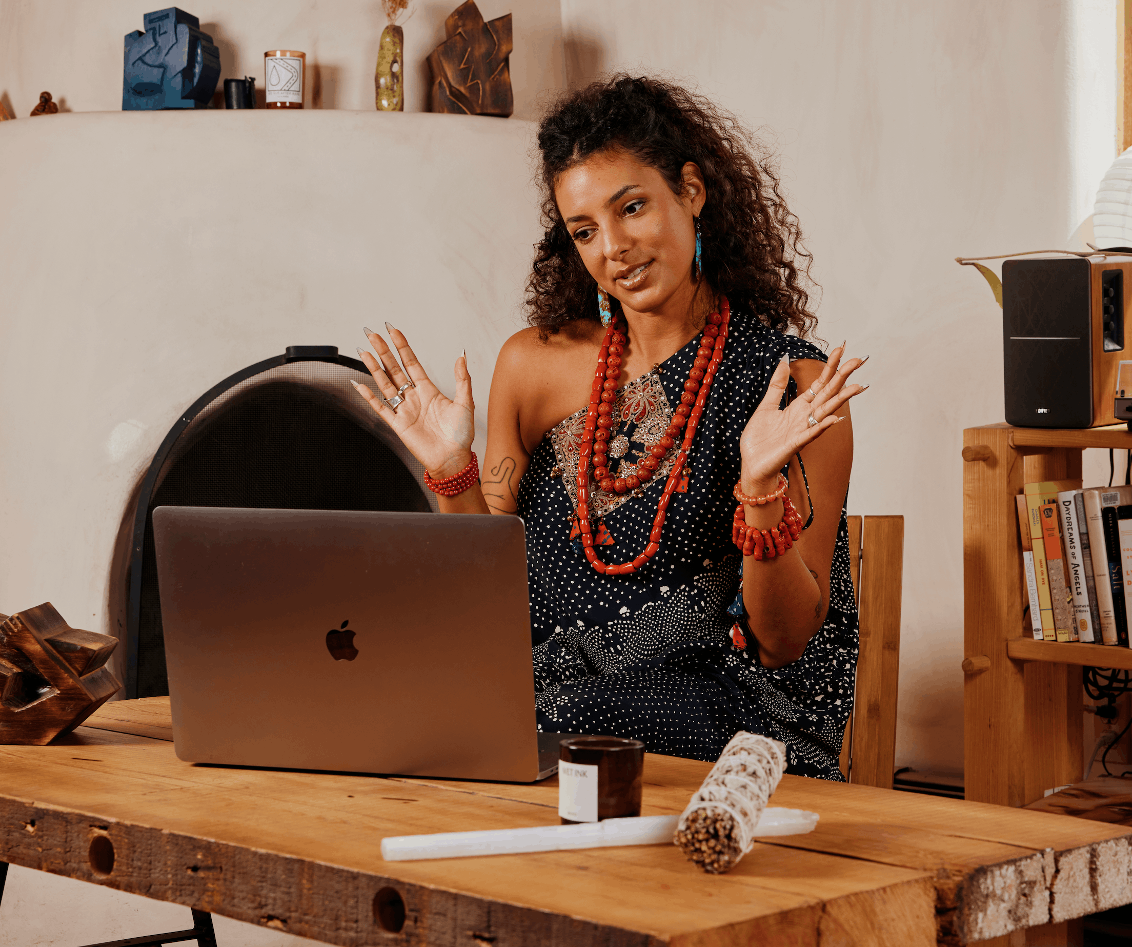 A woman with curly dark hair wearing colorful jewelry and a polka-dot dress is sitting at a wooden table with an iMac computer. Behind her, shelves display decorative items and books. A speaker and other objects are on the table.