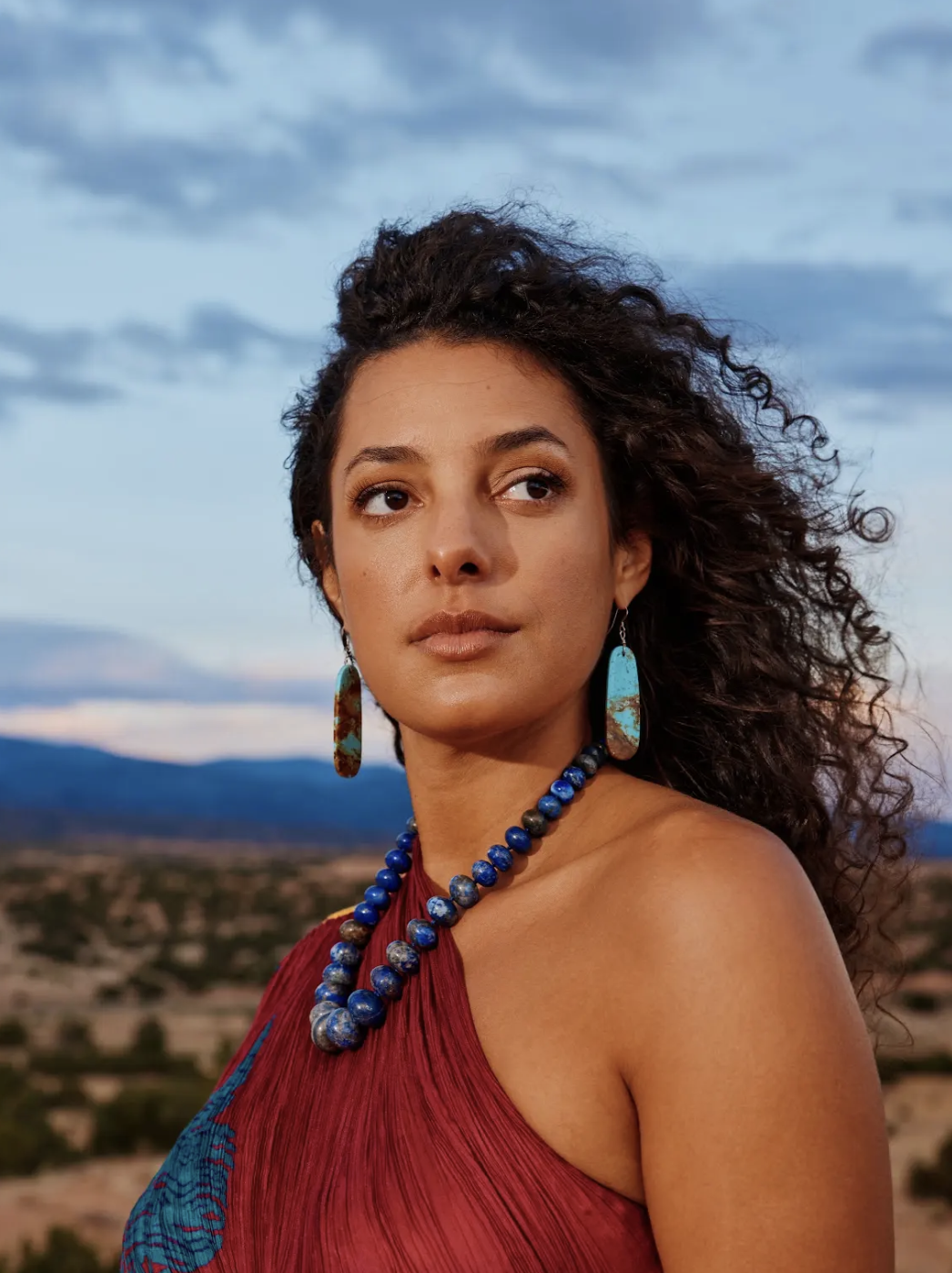 Portrait of a woman with curly hair wearing turquoise earrings and a blue beaded necklace, dressed in a red top, outdoors with a desert landscape and mountains in the background under a partly cloudy sky.