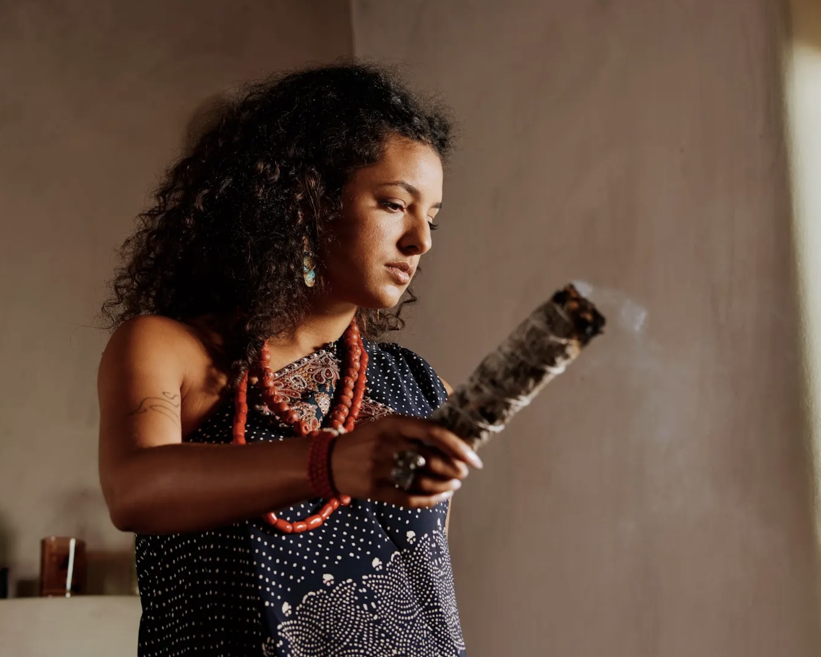 A woman with curly dark hair wearing traditional jewelry and clothing, holding a burning stick or torch, standing indoors against a plain wall.