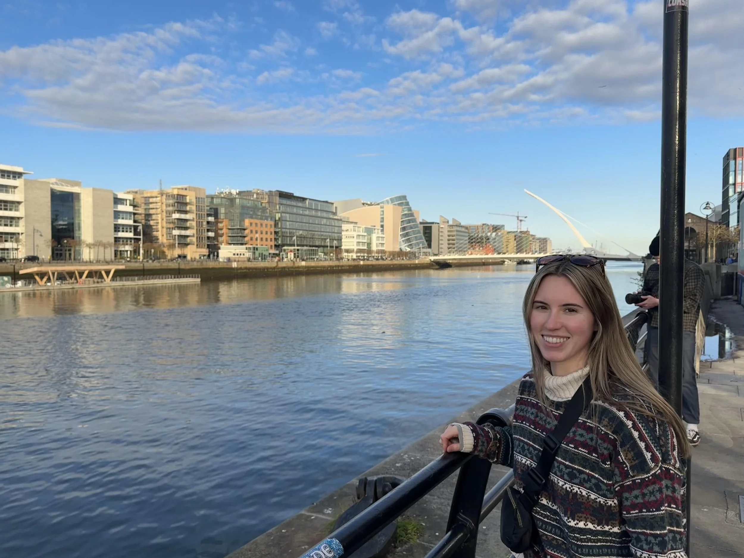 A young woman with long blonde hair smiling while leaning on a railing along a riverwalk, with a city skyline and blue sky with clouds in the background.