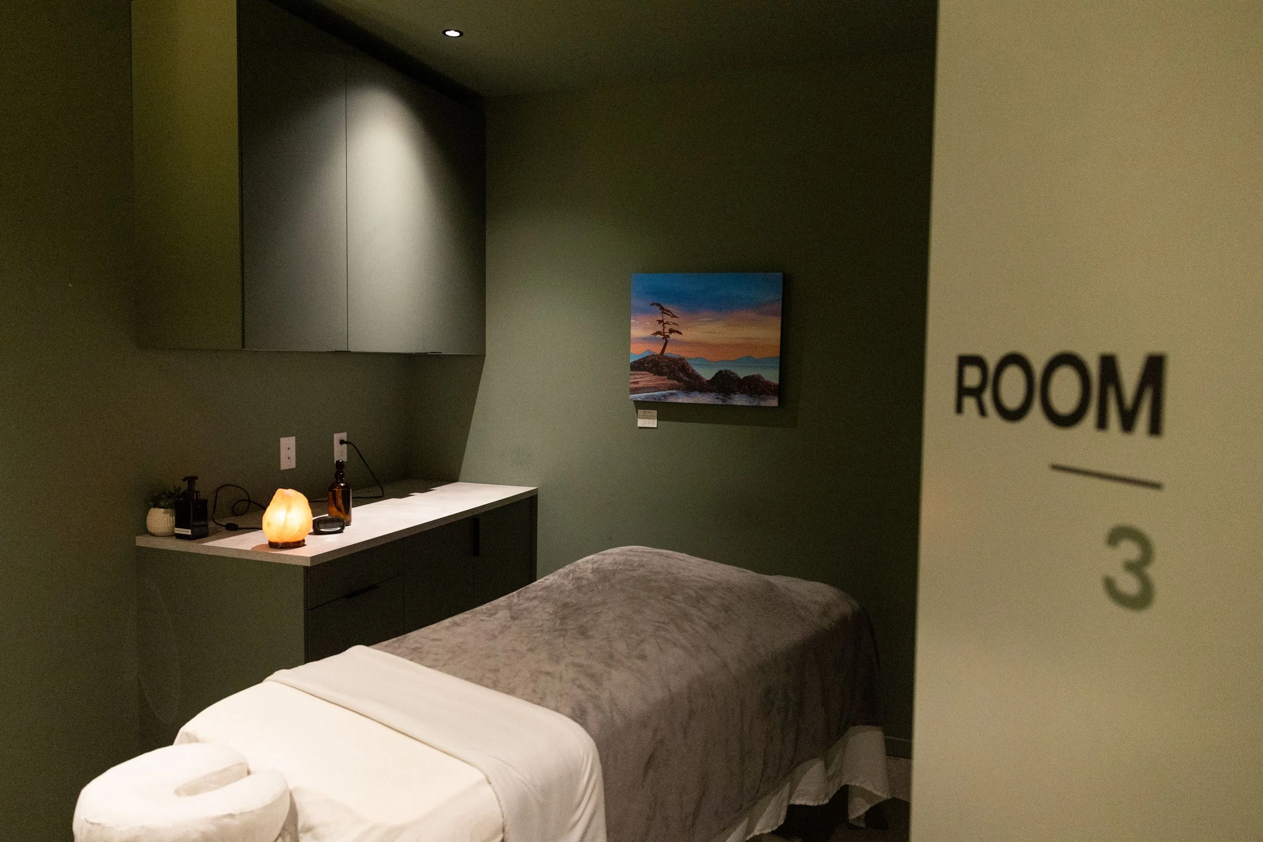 A hotel room with a bed, a nightstand with candles, a wall-mounted art piece of a landscape at sunset, and a salt lamp. The room has dark-colored walls and modern decor.