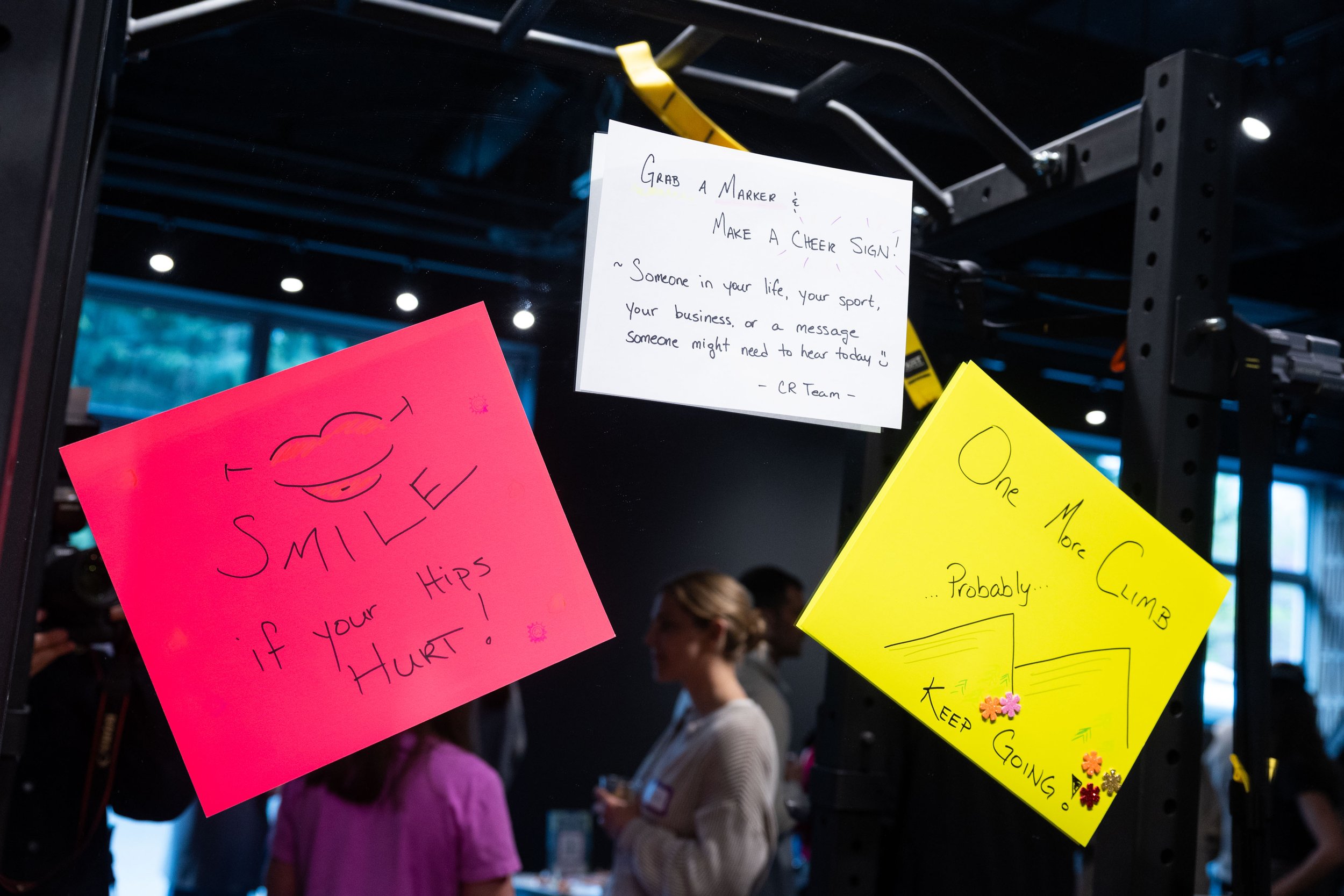 Colorful handwritten signs on a display shelf at an event. The visible signs include a white sign with motivational message, a pink sign with a smiley face and message, and a yellow sign with graphics and text about climbing. People are visible in the background, indicating a lively atmosphere.