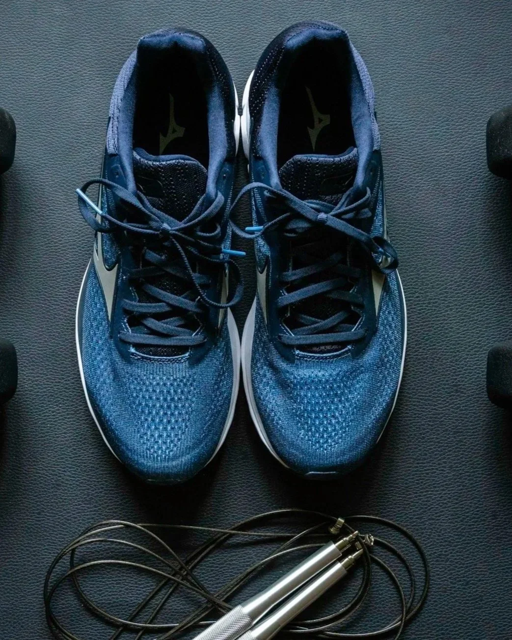 A pair of blue athletic running shoes on a black textured surface with a jump rope in front of them and a corner of a dumbbell on either side.