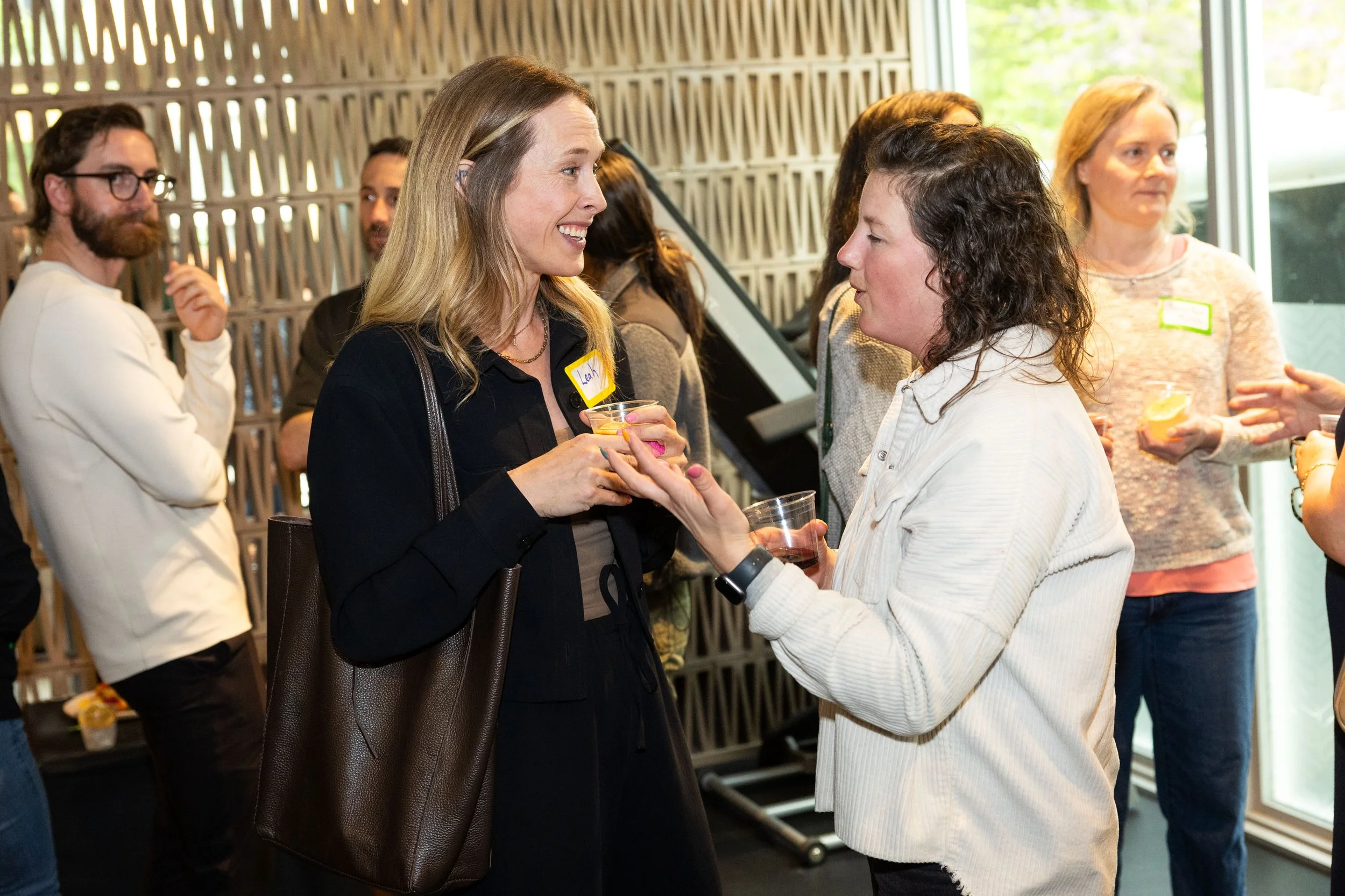 Two women engaged in a conversation at a social gathering, holding drinks, with other people in the background.