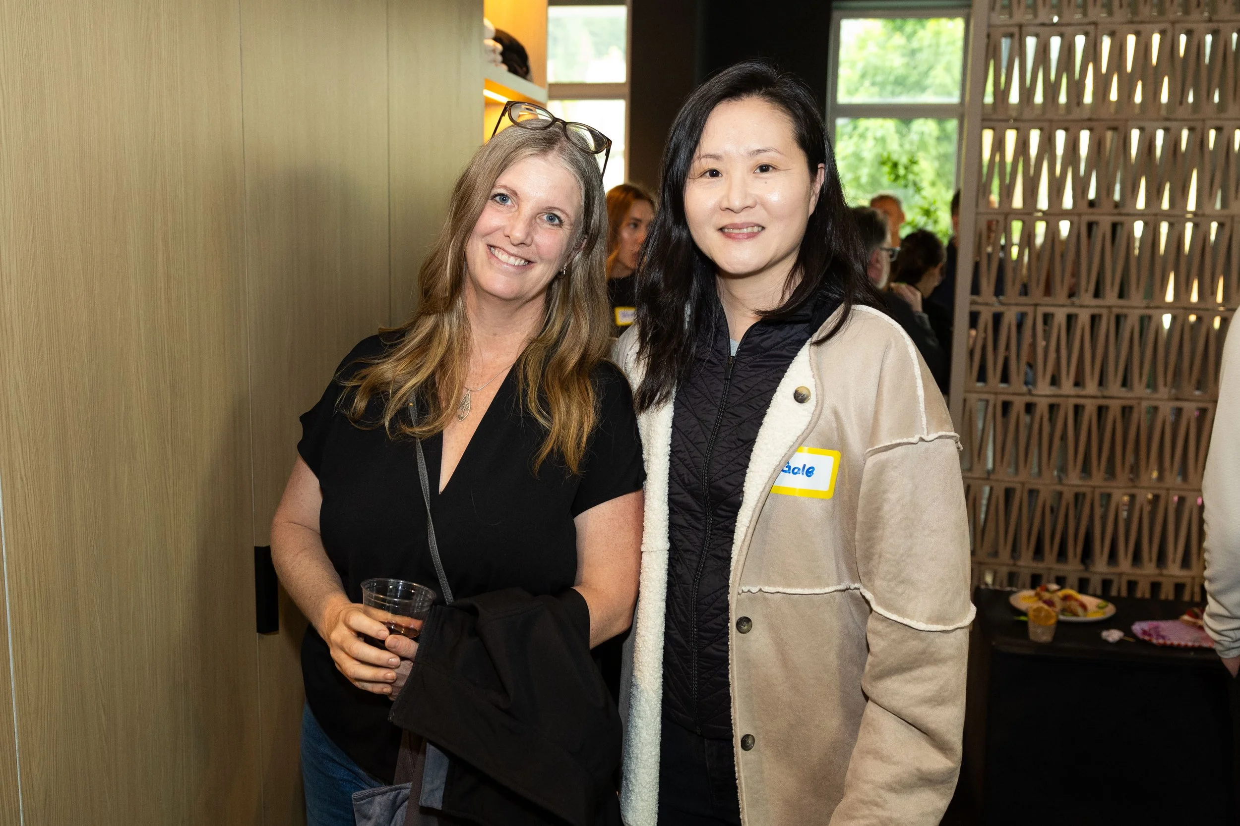 Two women posing for a photo at a social gathering, one holding a drink, with a background of people and a table with food.