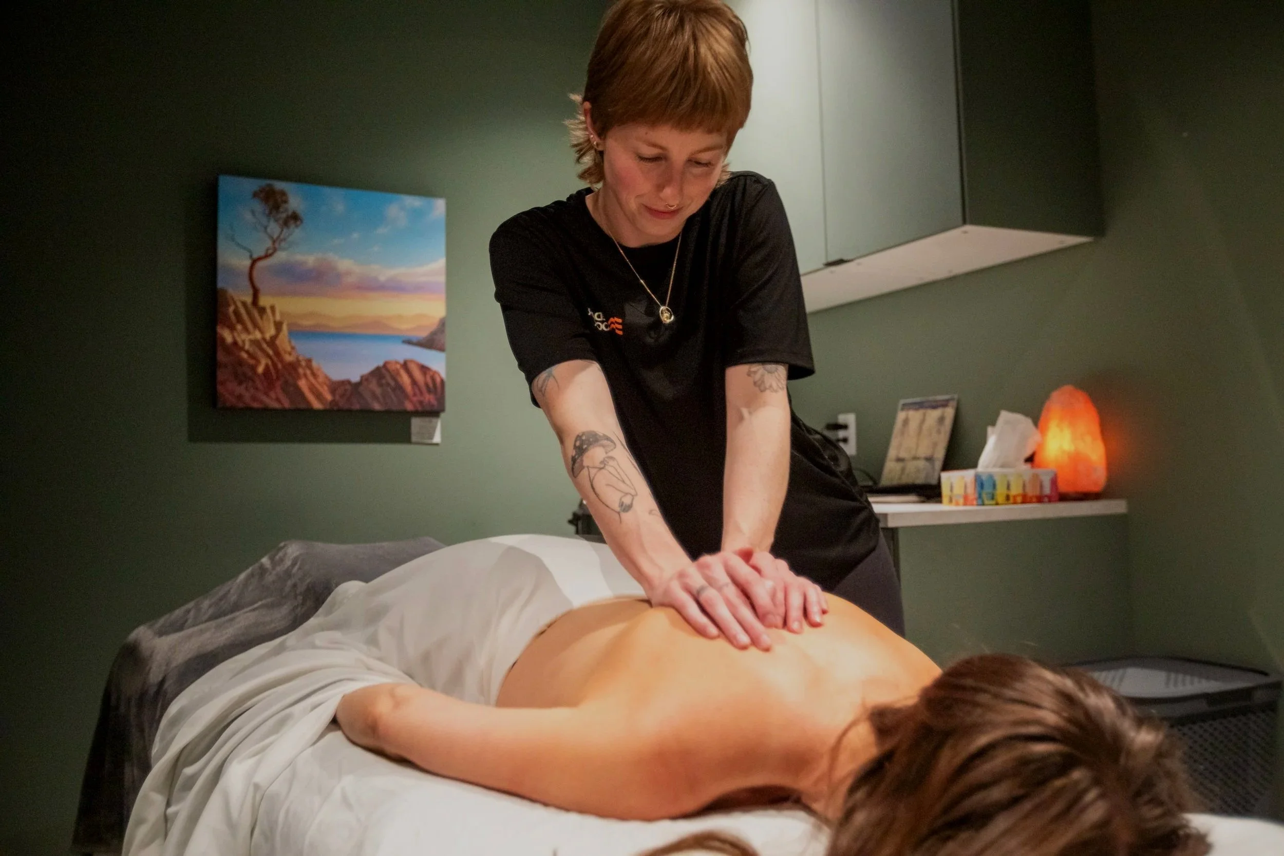 A woman with short hair and tattoos giving a massage to a person lying face down on a massage table in a room with green walls, colorful artwork, and a salt lamp.