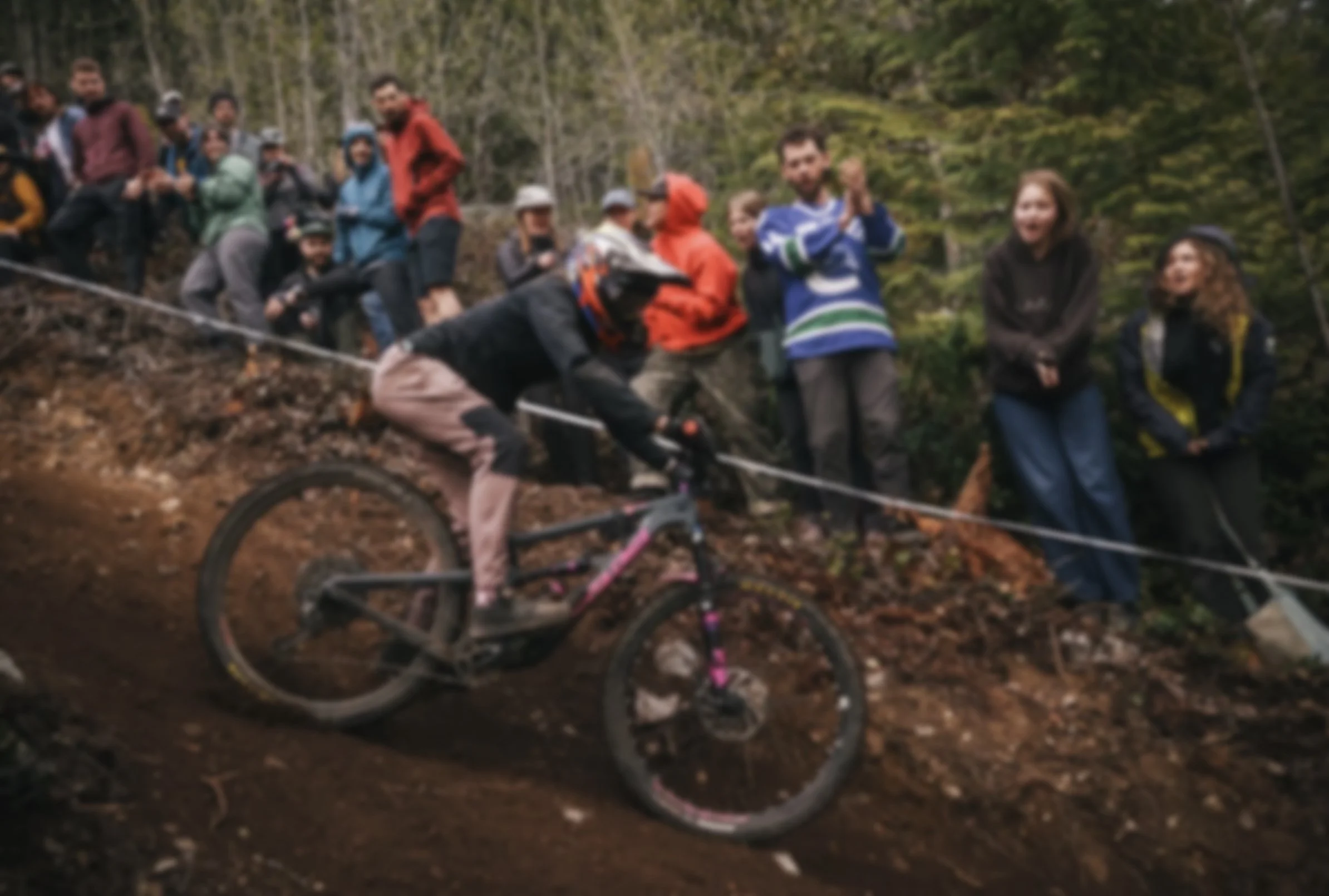 A group of people gathered outdoors in a wooded area, some standing and some sitting, with one person in the foreground riding a mountain bike on a dirt trail.