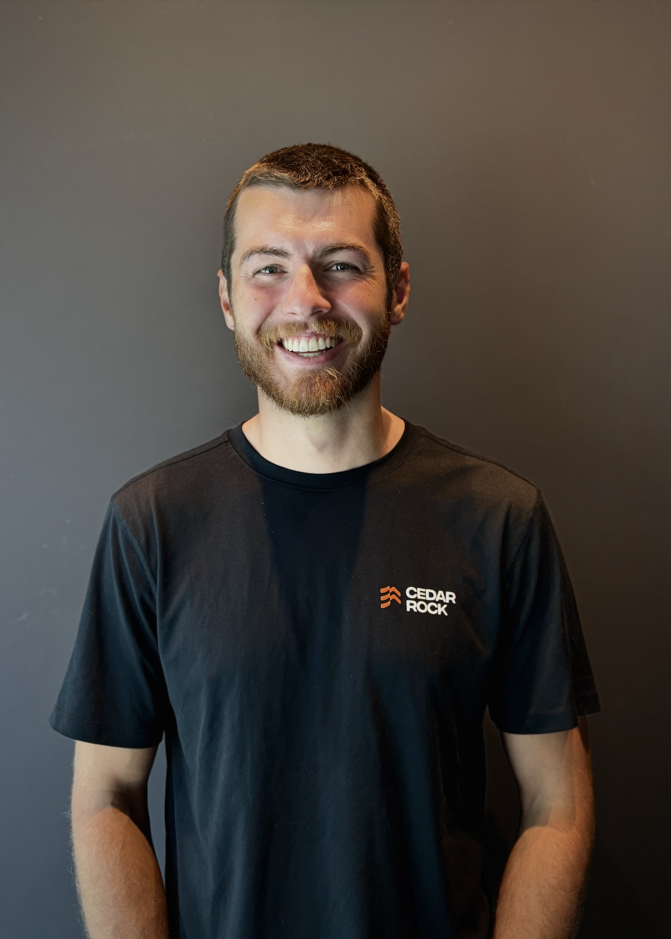 A young man with a beard and short hair, smiling, wearing a black Cedar Rock t-shirt, standing against a plain dark gray background.