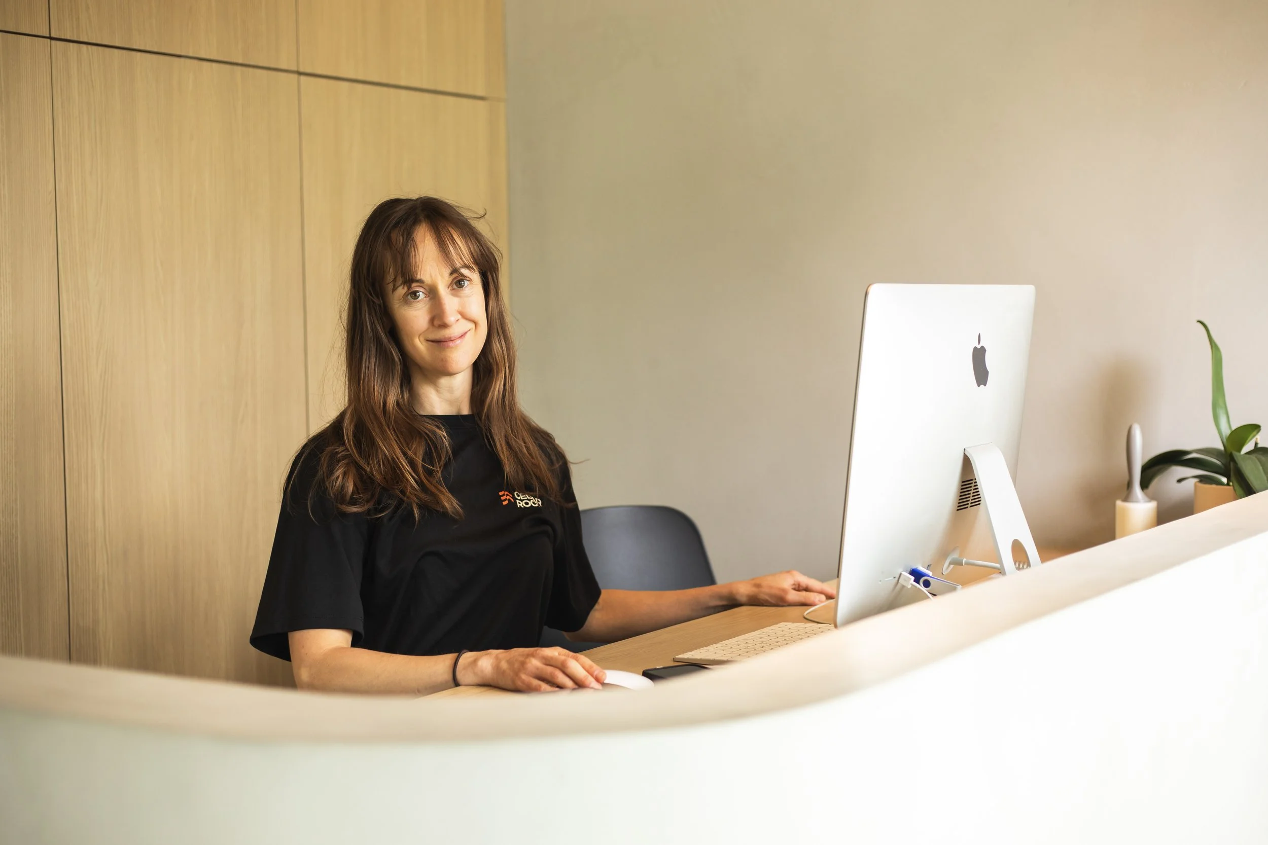 A woman with long brown hair sitting at a desk with a white Apple iMac computer in an office setting, smiling at the camera.