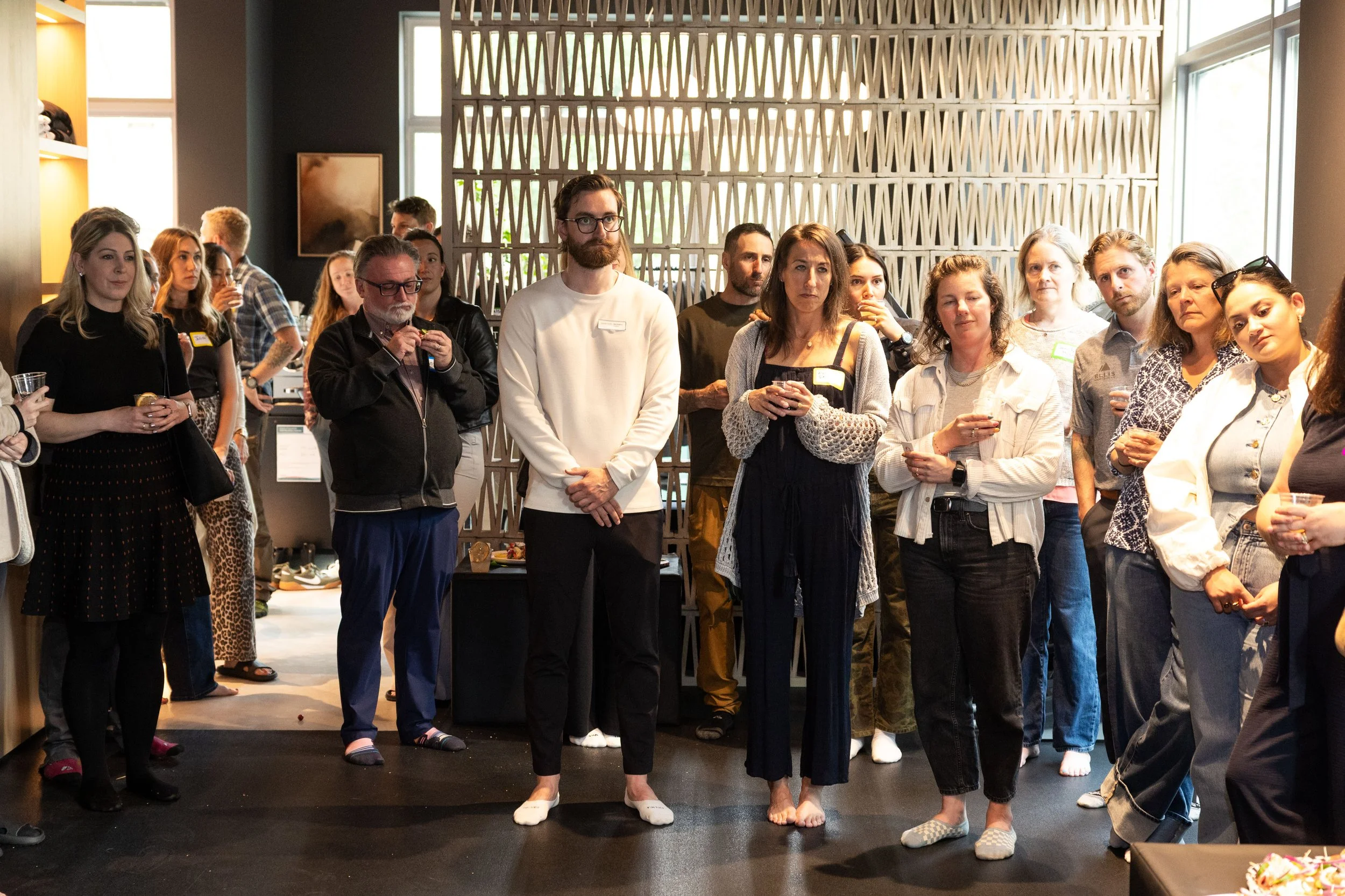 A diverse group of people attending an indoor event, some holding drinks, with most standing and listening attentively, near a decorative wall and large windows.
