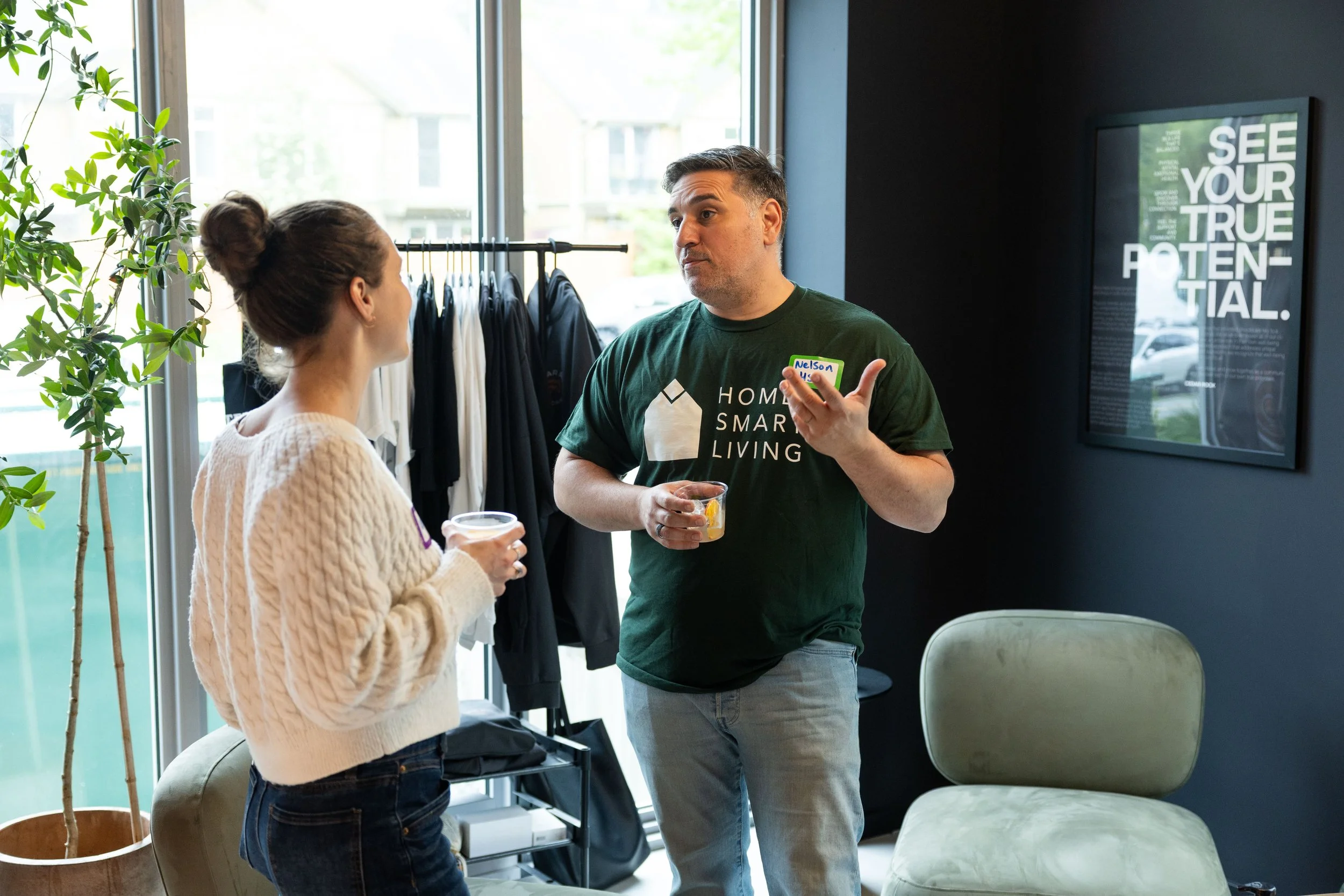 A woman and a man holding drinks and having a conversation indoors near a window and a black wall with a framed poster.