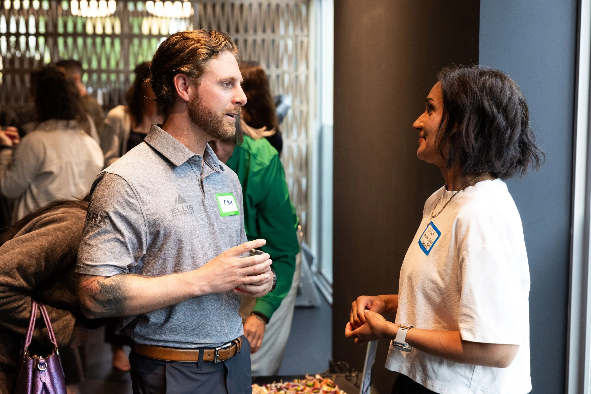 A man and a woman are engaged in conversation at a social event. The man is wearing a gray polo shirt and has a name tag that says 'CAM.' The woman is wearing a white top and has a name tag. They are standing near a table with food, and there are oth