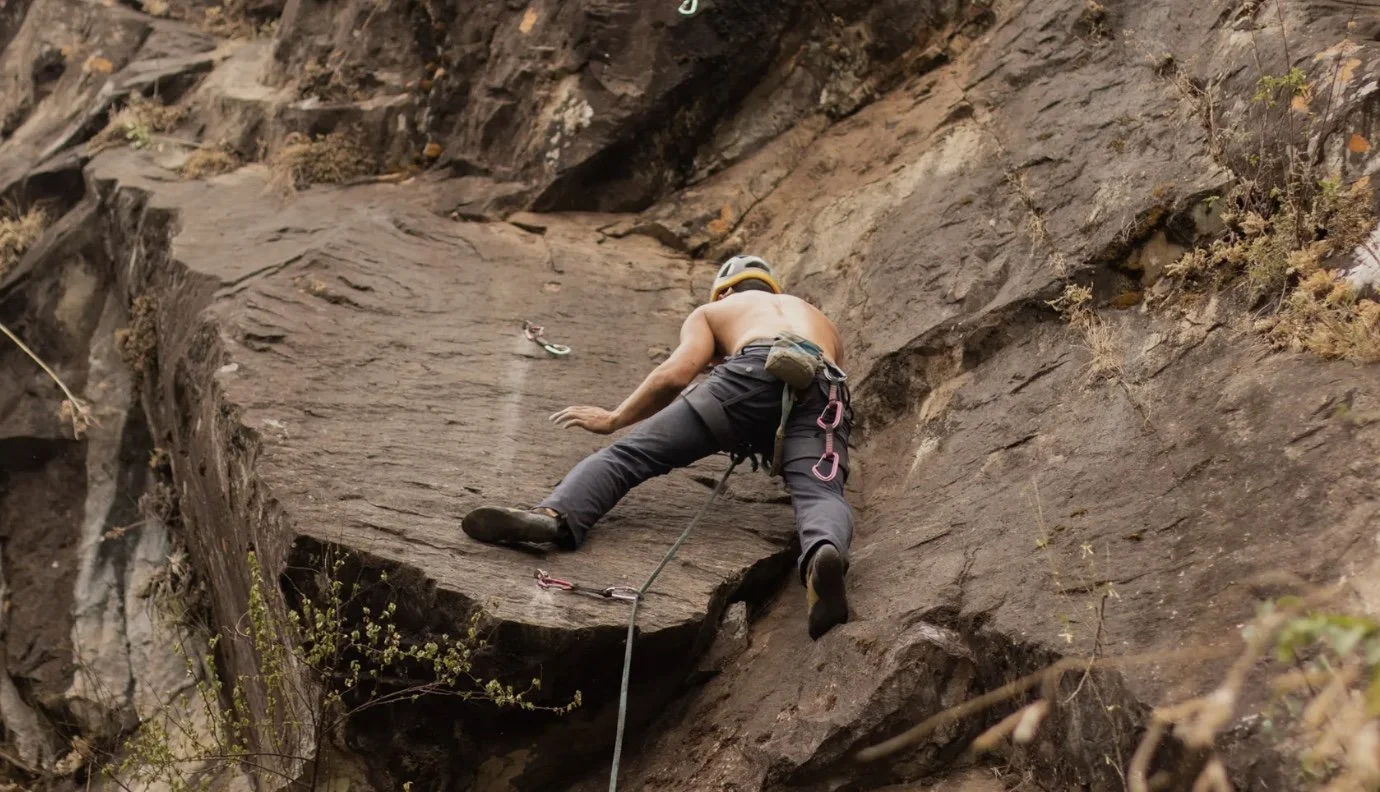 A shirtless male rock climber wearing a helmet climbing a vertical rock face with climbing gear.