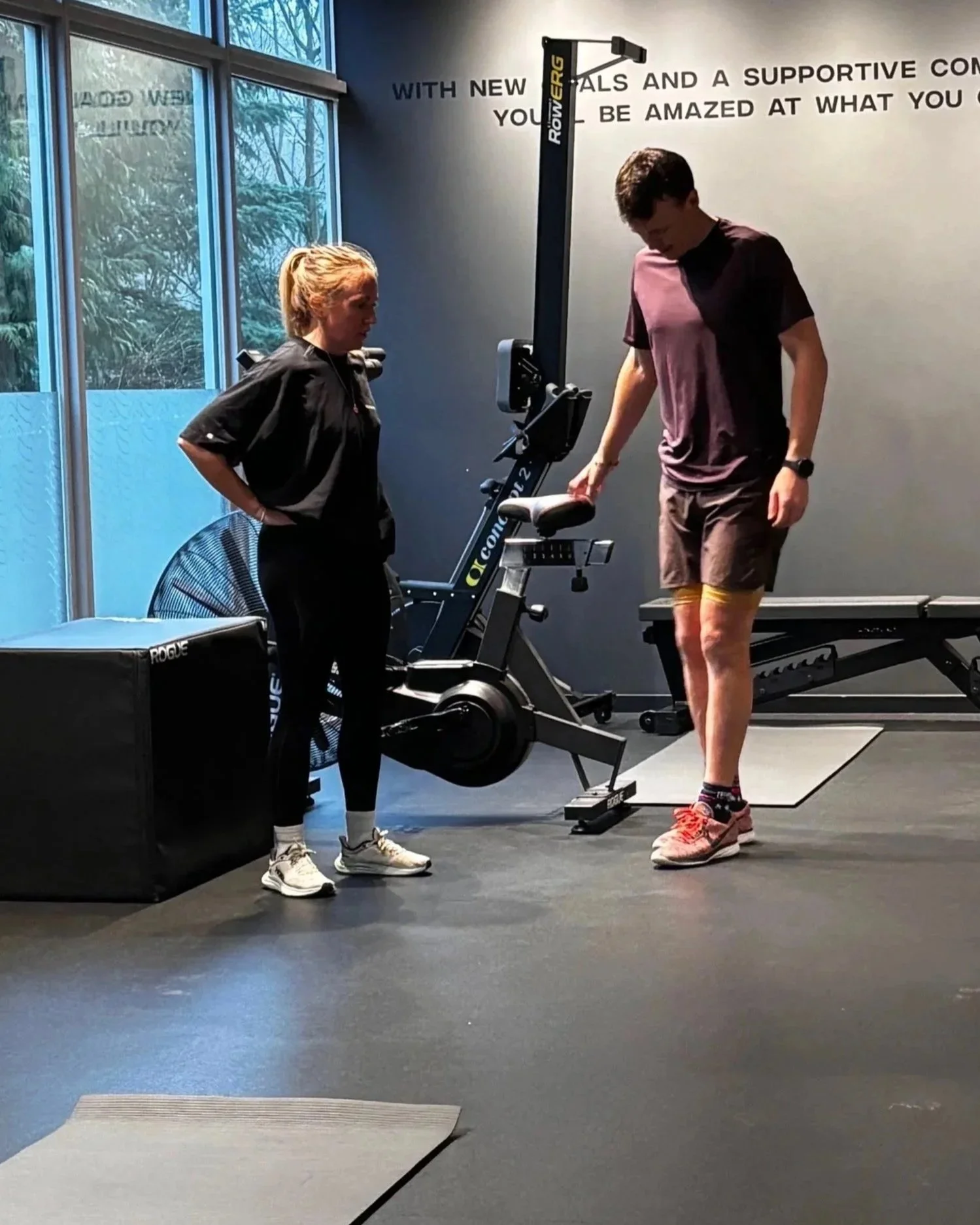 A person is standing on a exercise mat while a trainer or coach is looking at their form or position inside a gym. The gym has large windows and motivational text on the wall behind them.