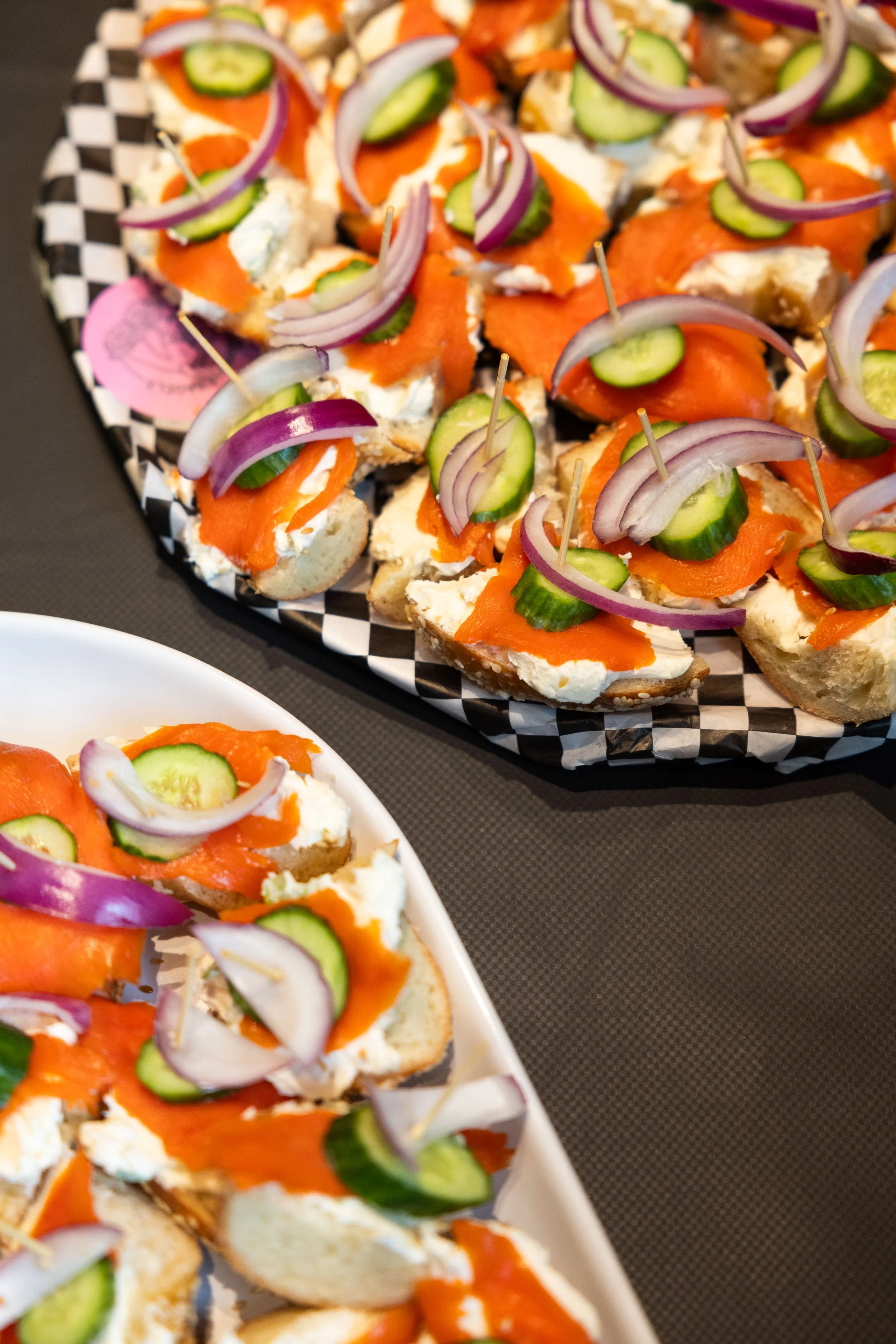 Assorted appetizers on a checkered black and white tray, topped with cucumber slices, red onion slices, smoked salmon, and a creamy spread.