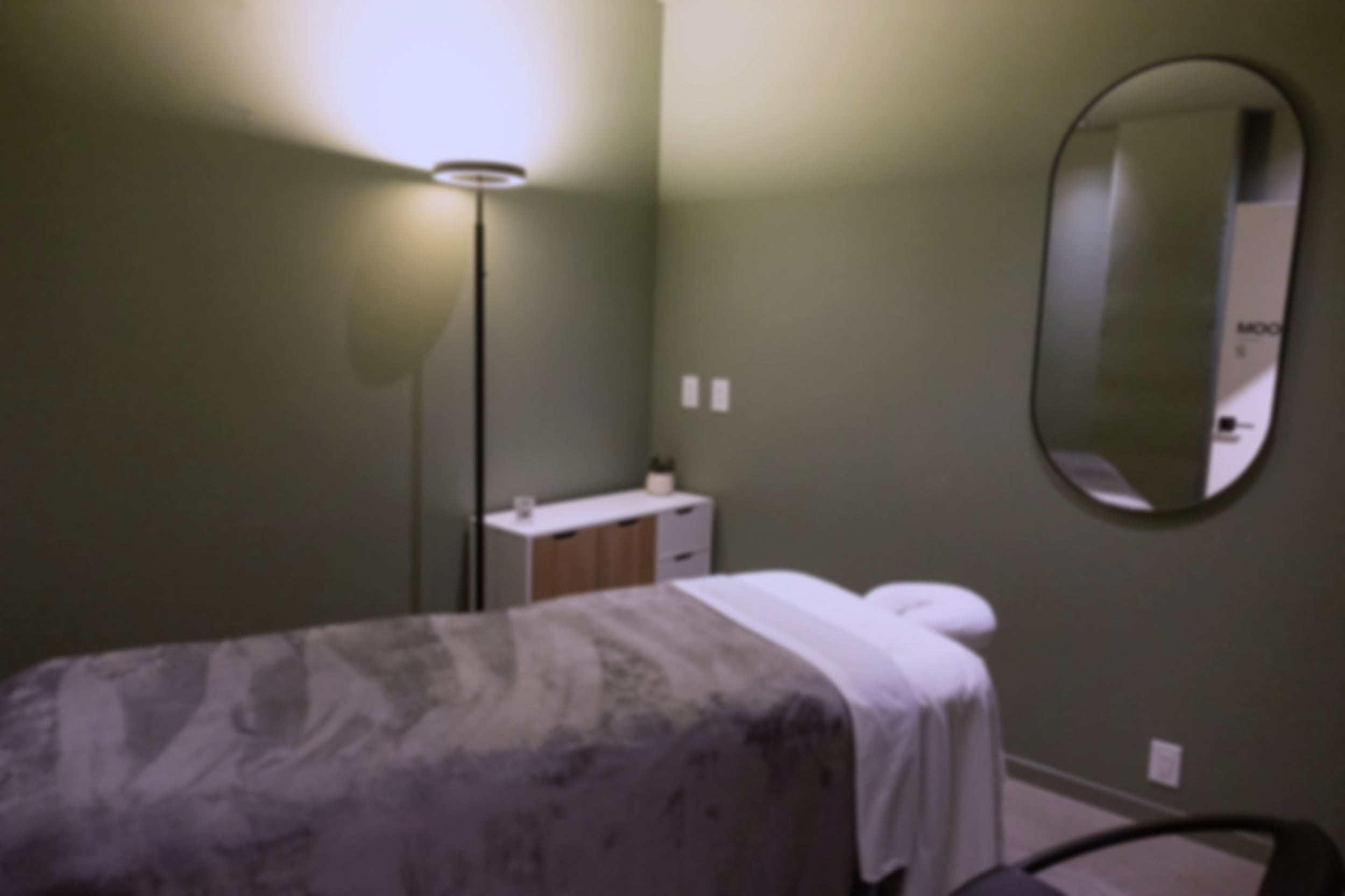 A massage therapy room with green walls, a bed with a gray and white patterned comforter, a bedside table, a mirror on the wall, a floor lamp, and a towel folded on the bed.
