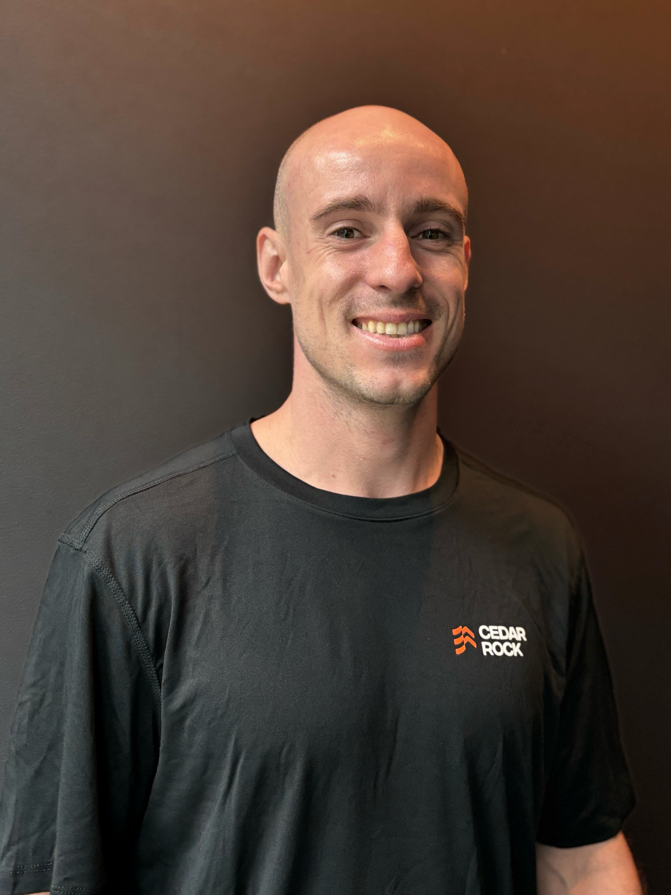 A man with a bald head and a broad smile, wearing a black athletic t-shirt with the Cedar Rock logo, standing against a dark wall.