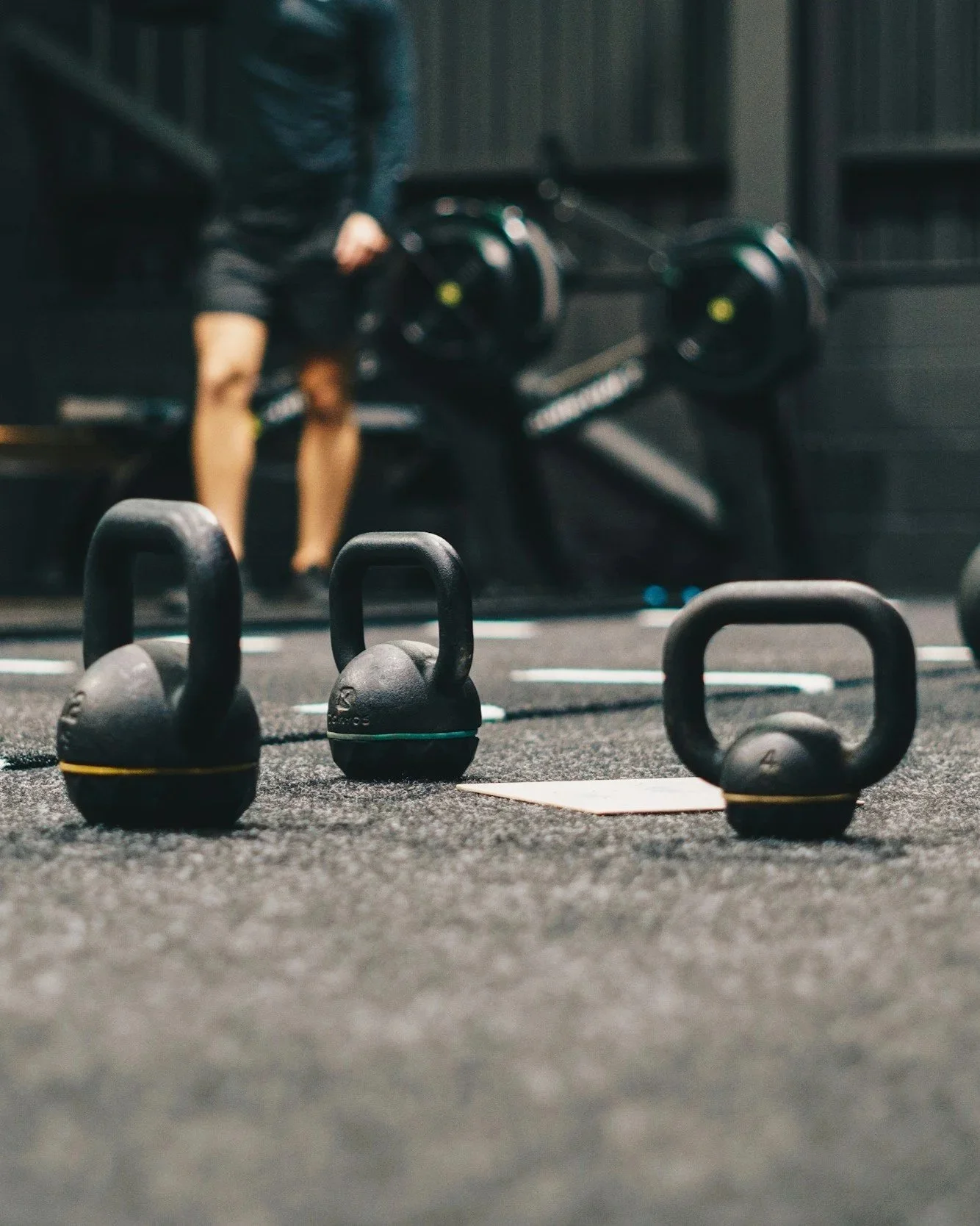 Several black kettlebells on a gym floor with a person in workout clothes in the background near exercise equipment.