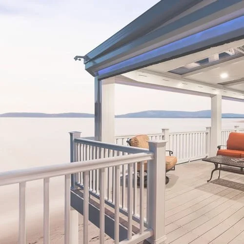 Lakeside deck with white railings and outdoor seating, including a cushioned chair and sofa, under a covered structure with a scenic waterfront view.