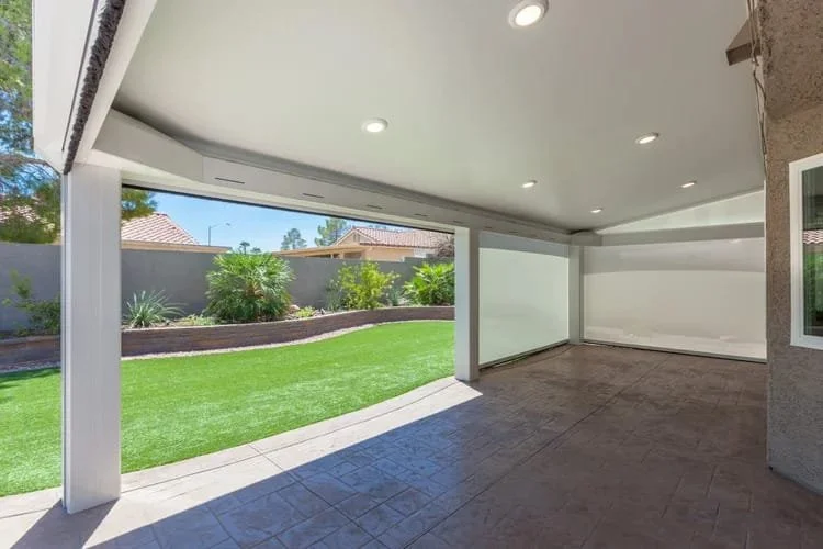 Covered patio with retractable screens, stamped concrete flooring, and view of a landscaped backyard with artificial grass and plants.