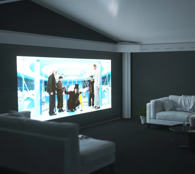 Modern living room with a large screen displaying a scene with several people. The room is dimly lit with gray couches and a small round table.