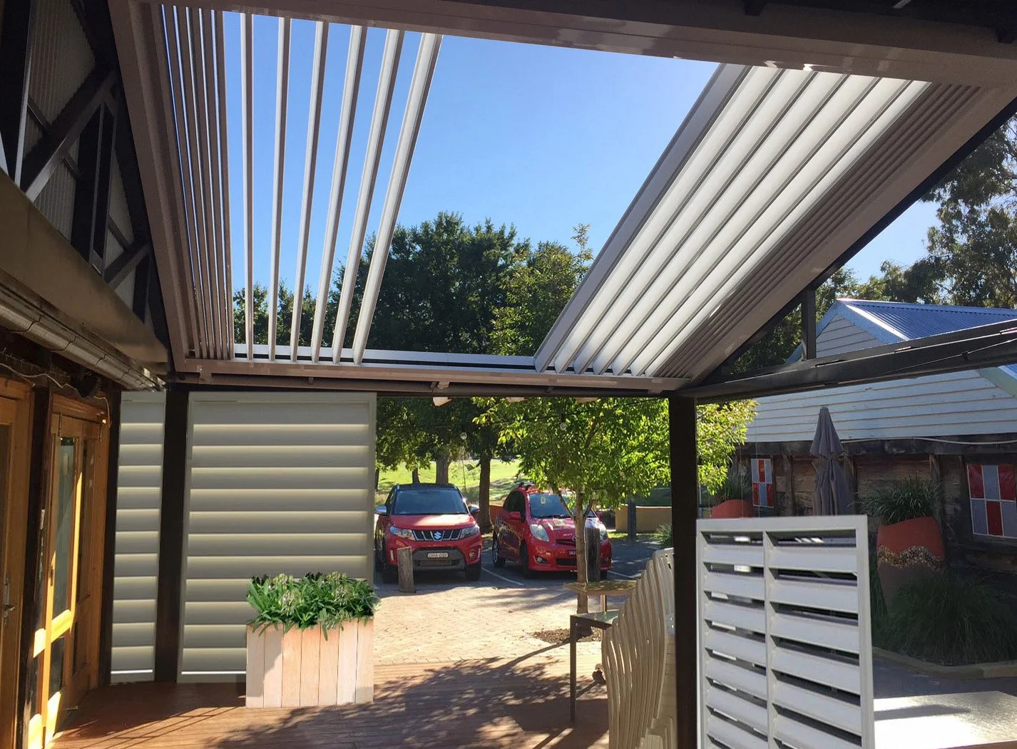 Outdoor patio area with a retractable roof, featuring slatted panels. The area includes wooden framed glass doors and a planter with greenery. Two parked red cars and trees are visible in the background.