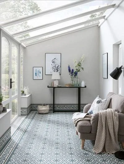 Bright sunroom with floor-to-ceiling windows, gray sofa, patterned tile flooring, minimalist artwork, and plants.