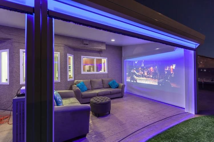 Outdoor home theater with a large projection screen displaying a movie scene, surrounded by a gray sectional sofa with teal pillows, under ambient blue lighting in a covered patio setting.