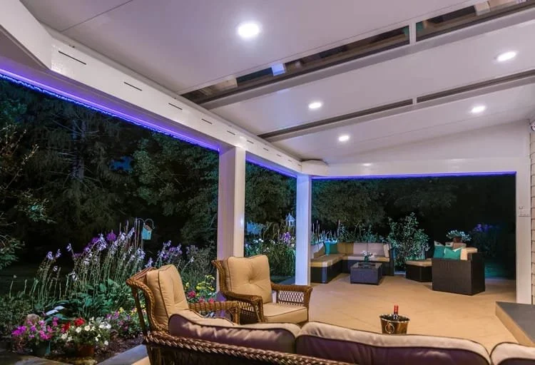 Outdoor patio with wicker furniture and cushions, surrounded by a garden with flowers and trees, featuring overhead recessed lighting and blue LED strip lighting at night.